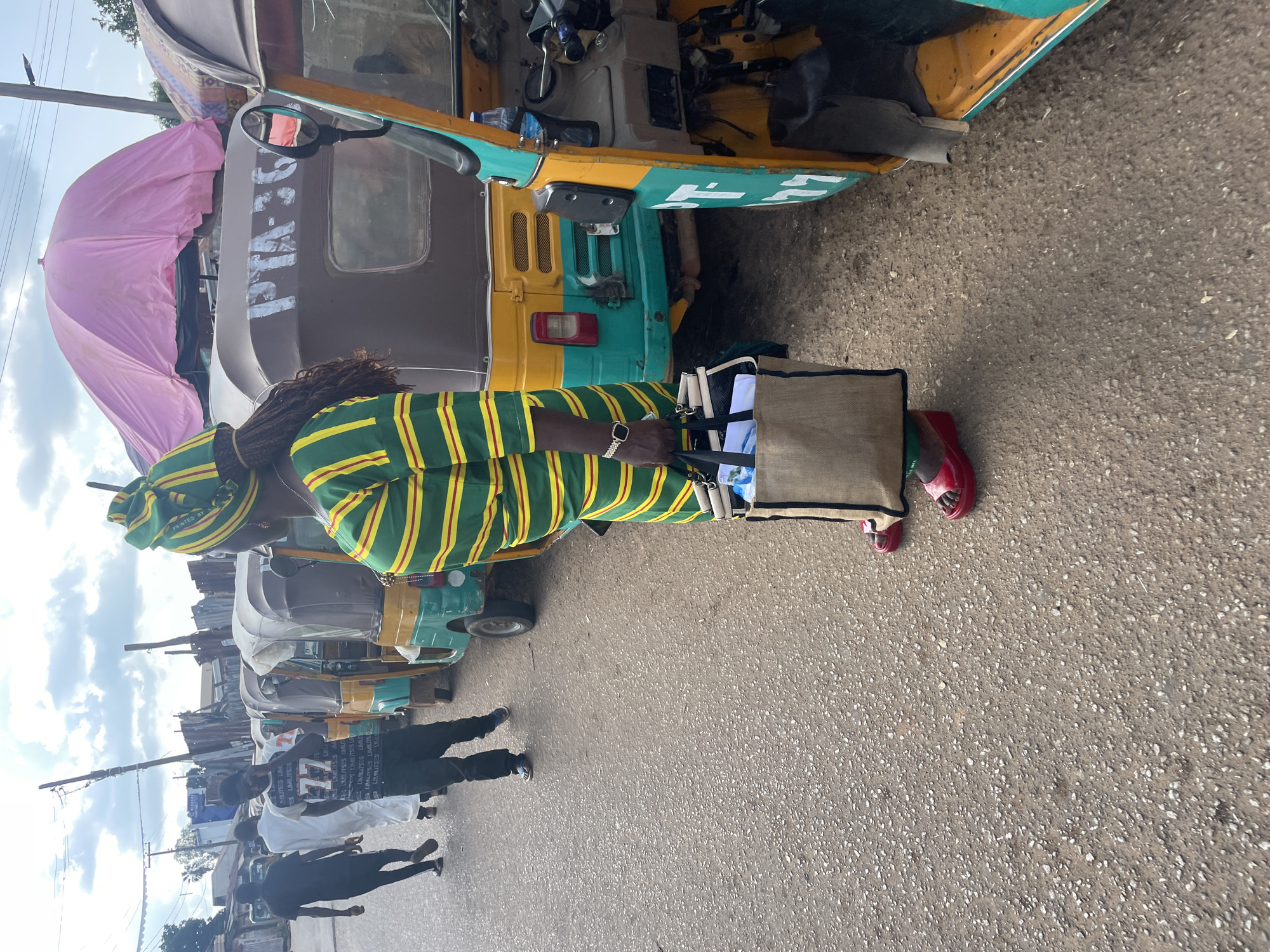 The image depicts a street scene in Gura Top, Nigeria, with a focus on a woman dressed in a green and yellow striped outfit and headscarf. She is standing to the right of the frame, holding a large canvas tote bag. Her feet are clad in red wedge sandals. Behind her, several auto-rickshaws, locally known as "Keke Napeps," are parked along the roadside. One of the Keke Napeps has the painted inscription "PTA-367" on its side. In the background, other individuals are walking along the street, and there are various structures and makeshift stalls, suggesting a market or busy thoroughfare. The sky is partly cloudy. The road surface is asphalt with visible aggregate.