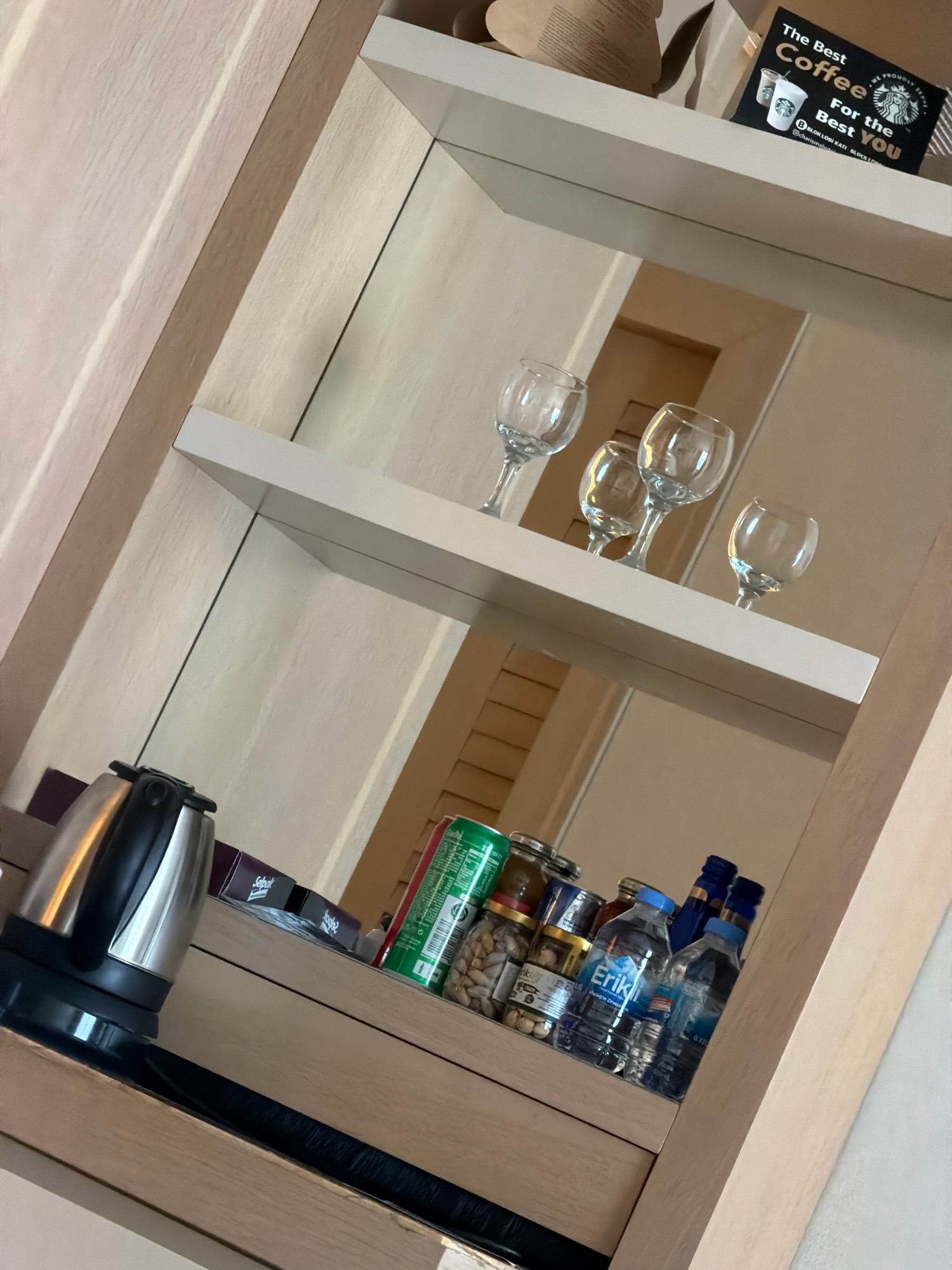 The image shows a hotel room minibar. On the top shelf, there is a Starbucks promotional card, a coffee cup, and what appears to be a straw. On the middle shelf, there are four wine glasses. On the bottom shelf, there is an electric kettle, a box of what might be tea or coffee, a can of green soda, a jar of nuts, a small bottle of alcohol, and two bottles of water. The minibar is situated within a wooden cabinet with mirrored backing. The overall scene suggests a hotel room setting in Kuşadası, Turkey, given the context.