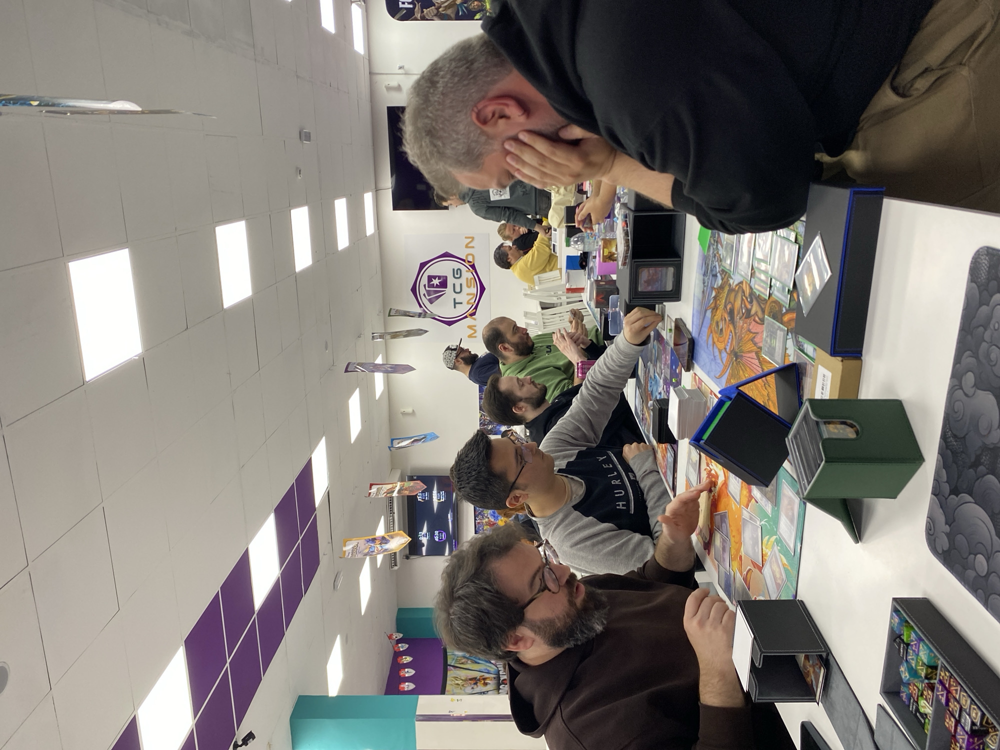 The image shows a group of people seated at a long table in what appears to be a game store or convention setting in Gijón/Xixón, Spain. They are engaged in a tabletop game, possibly a collectible card game like Magic: The Gathering, as cards are spread out on playmats in front of them. The table is white. Various accessories, such as card boxes and dice, are also visible on the table. Overhead are rectangular ceiling lights arranged in a grid pattern. The walls are white with a purple border running along the top. A logo with the word "MANSON" is present on the wall. Banners and posters are hanging from the ceiling.
