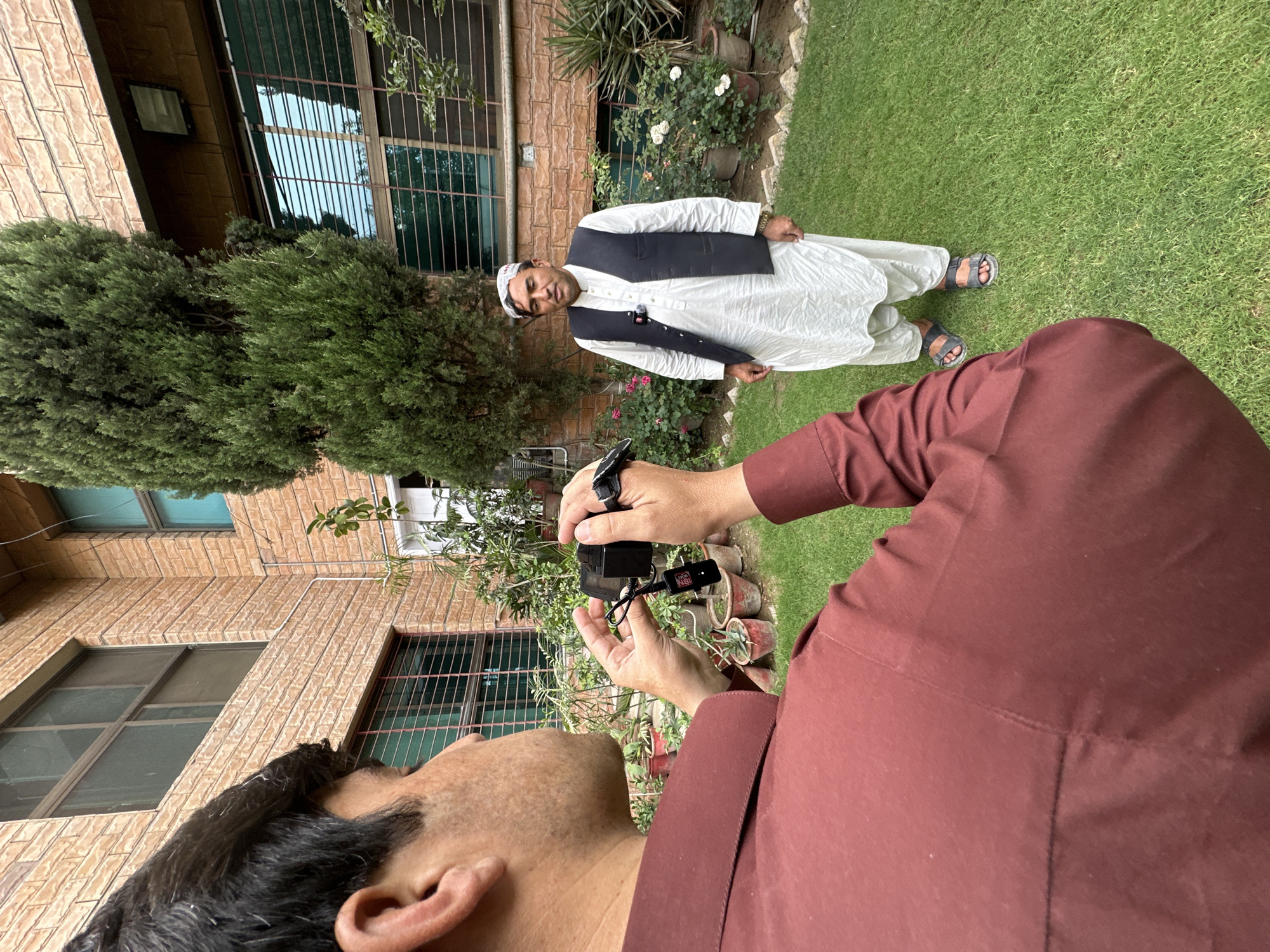 The image captures a scene in Quetta City Tehsil, Pakistan, featuring two men. One man stands in the background, dressed in traditional Pakistani attire consisting of a white salwar kameez and a black waistcoat, with sandals on his feet. He poses near a building with barred windows and potted plants. The other man is shown from a lower perspective, close to the camera. He wears a maroon-colored shirt and appears to be holding a camera, suggesting he is taking a photograph of the first man. The environment is lush, with green grass and vegetation surrounding the individuals and building. The buildings in the background suggest a residential area.