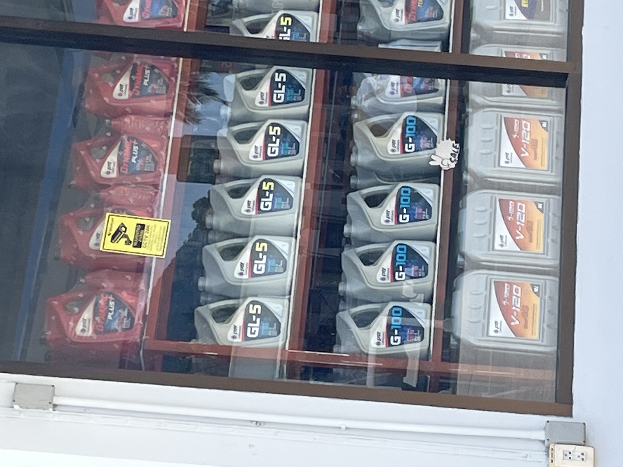 The image shows a display window of a store, likely a mechanic shop or auto parts store, in Koh Phangan, Thailand. The window displays several shelves filled with various motor oil containers. The products are predominantly red and silver-colored plastic jugs with labels indicating different types of oil, such as "DYNAMIC PLUS", "GL-5", "G-100", and "V-120". Some of the bottles have labels with the "PTT Lubricants" logo. A yellow sticker with a CCTV camera icon and text "CCTV 24h" is visible on the top shelf. A small "SALE" sign with a hand gesture is placed on the middle shelf. The display is illuminated from within, and the reflection of trees and sky is visible on the glass, indicating an outdoor setting. The exterior of the building appears to have white siding and a white electrical conduit with a beige electrical outlet.