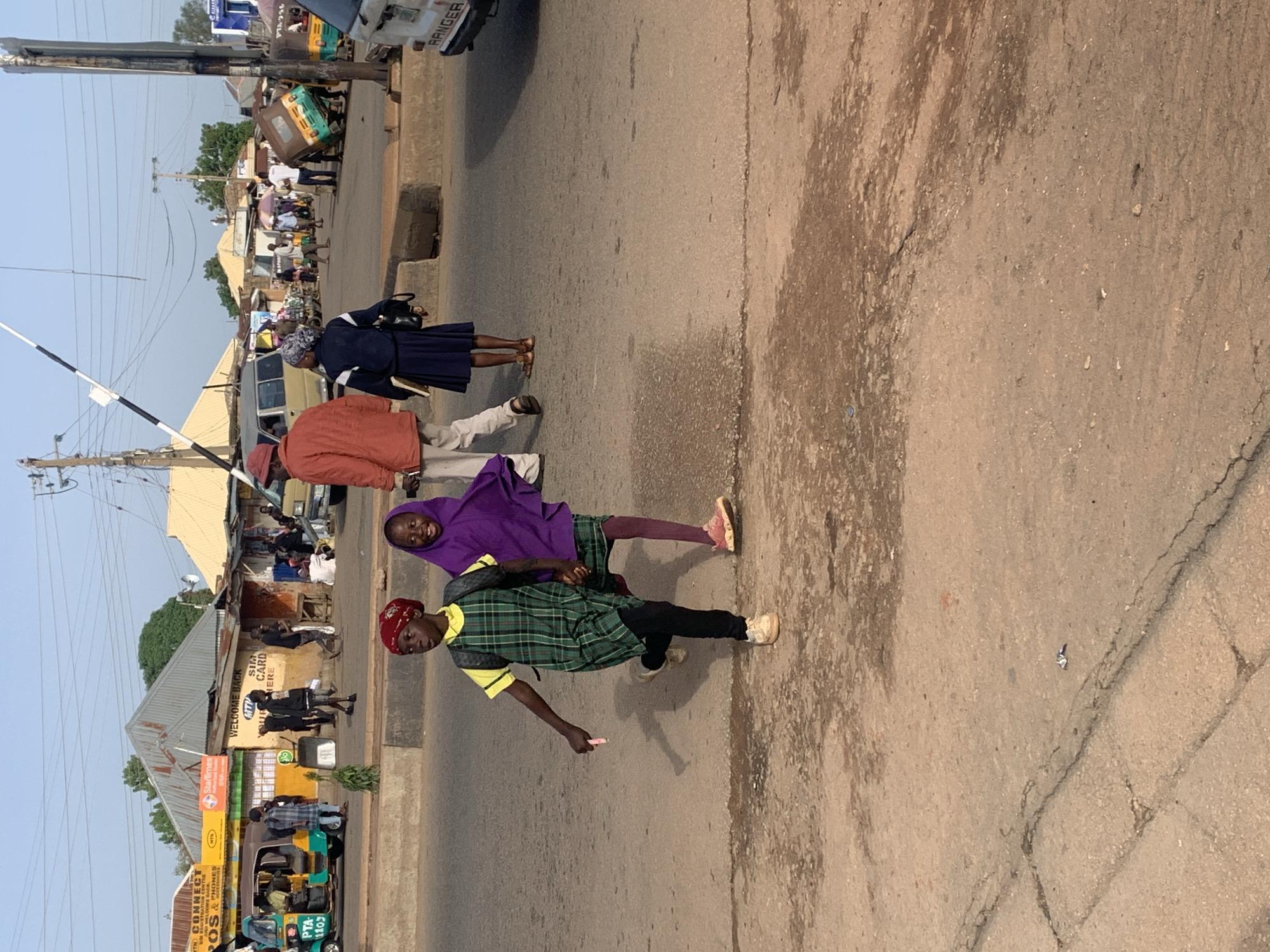 This image captures a street scene in Gura Top, Nigeria. The primary subjects are several individuals, including two children in the foreground. One child, wearing a purple hijab and pink leggings, is being playfully embraced by another child dressed in a green and yellow plaid shirt and black trousers. Both children appear to be smiling. Behind them, an adult wearing an orange jacket and a woman in a dark blue uniform with a headscarf are also visible.
The setting is a busy street with vehicles, including a tricycle, parked and driving. Buildings line the street, suggesting a commercial or residential area. One building has signage that includes "MTN CONNECT," "POS & PHONES," "WELCOME BACK," and "SIM CARD HERE." Other visible text includes "PTA-256" and "PTA-1103" on a vehicle.
The lighting suggests it is daytime, possibly late afternoon given the shadows. The sky is clear. The overall mood appears to be lively and indicative of daily life in a community. The presence of the tricycle and the various storefronts point to a functioning local economy.
