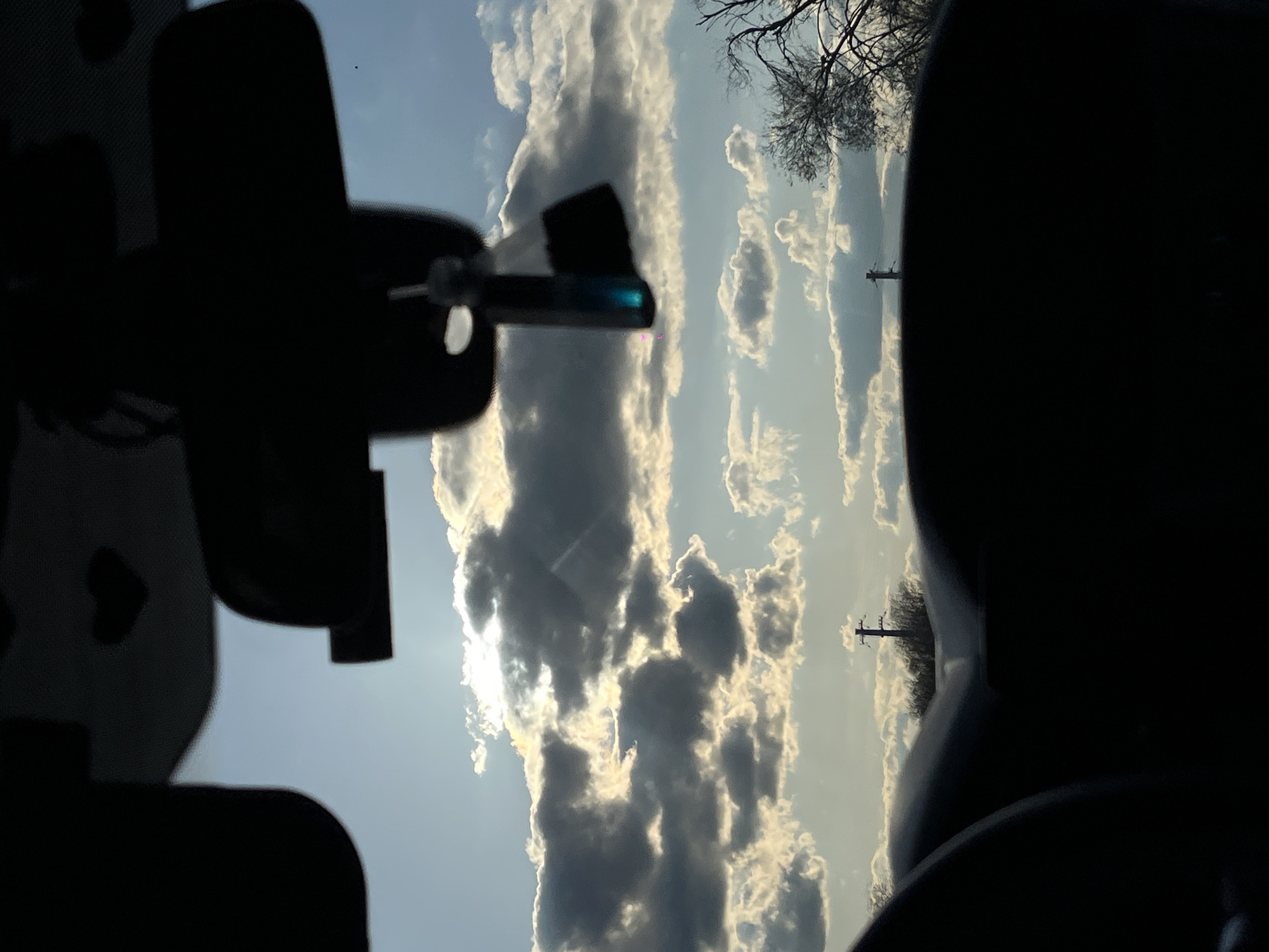 The media file, originally oriented vertically, displays a landscape view from within a vehicle, looking out at a partially cloudy sky over Ternovskaya, Russia.
In the immediate foreground, dark, indistinct silhouettes of the car's interior frame the top, left, and bottom-right of the image. On the left side, what appears to be the rearview mirror assembly is visible in silhouette, with a small, modern device attached to it, exhibiting a distinct blue-green reflection. On the bottom-right, another dark silhouette suggests the top of a car seat or part of the dashboard.
The central and right portions of the image are dominated by a bright sky filled with textured cumulus clouds. Sunlight intensely backlights these clouds, creating dramatic contrasts of bright white highlights and darker, shadowed areas. The sun itself is partially obscured but its strong presence is evident. Against this bright sky, faint, dark, bare tree branches are visible on the far right. Further into the distance, two slender utility poles with crossbars stand vertically, silhouetted against the bright clouds.
The scene depicts daytime, likely early morning or late afternoon, given the intense but soft quality of the sun filtering through the clouds. The weather is partly cloudy. The presence of utility poles and bare tree branches suggests a rural or semi-rural setting during a colder season like winter or early spring. No visible text is present in the image.