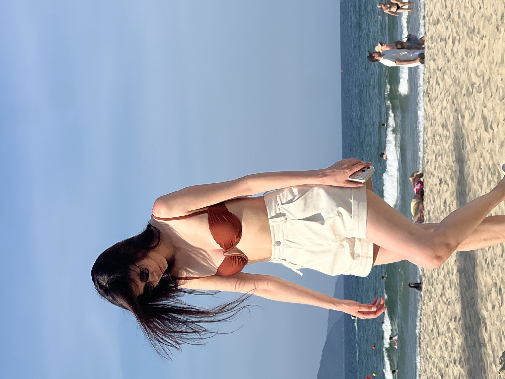 A young woman with long, dark hair and sunglasses walks along a sandy beach in Da Nang, Vietnam. She is wearing a reddish-brown bikini top and cream-colored shorts. Her hair is blowing in the wind. In the background, the turquoise ocean meets the horizon under a clear blue sky. Waves are crashing on the shore, and a few people are visible swimming and relaxing in the water and on the beach. The coastline is lined with low, green hills. The overall atmosphere is serene and summery.