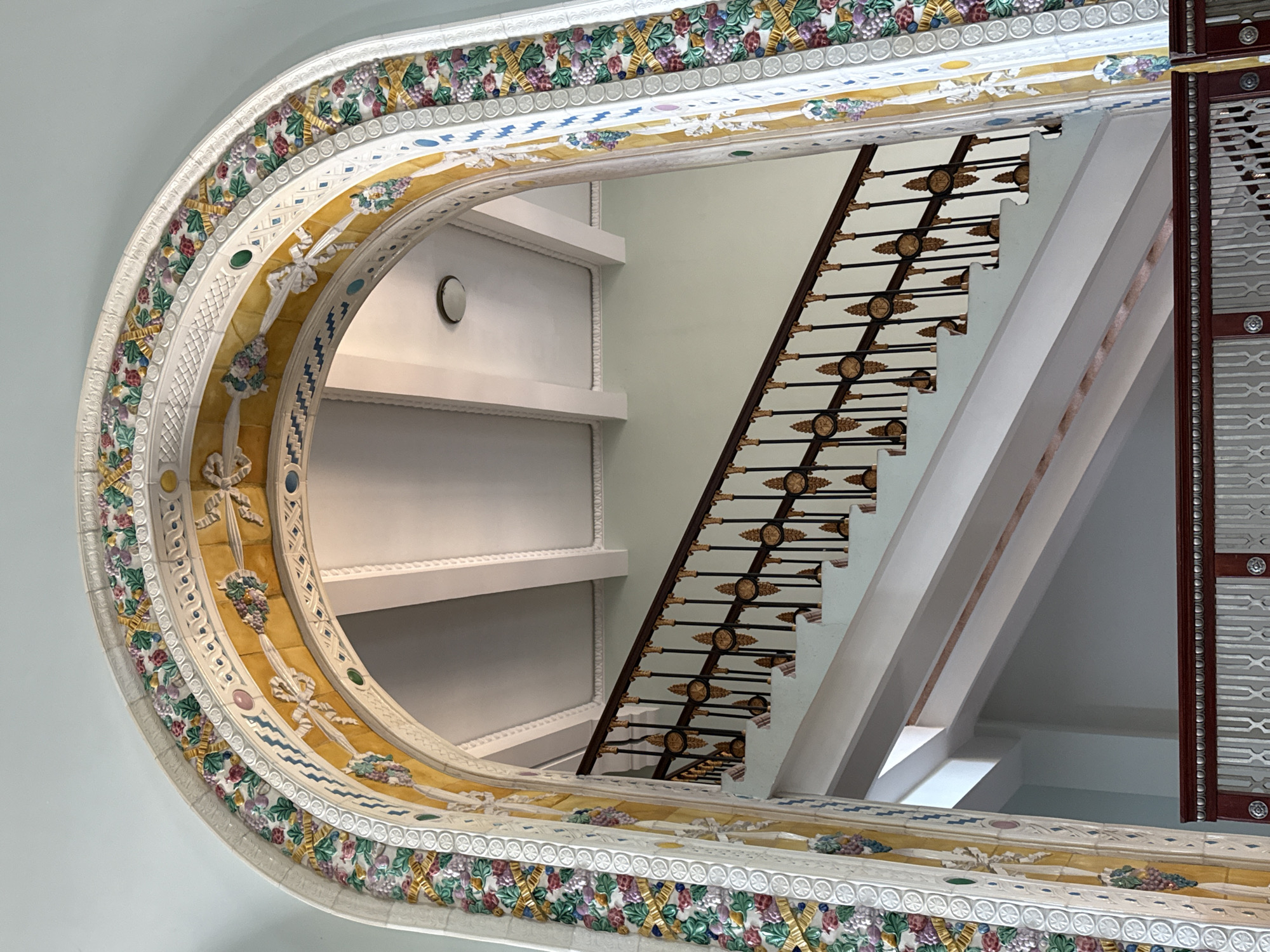 The image displays an upward-angled view through a highly ornate archway, revealing a grand staircase and interior architecture beyond. This scene is located within the Sanatorium Ordzhonikidze in Sochi, Russia. The archway is meticulously decorated with multi-colored ceramic or tiled elements. Its outer band is white with a repeating pattern of small circles. Inside this, a prominent relief band features green leaves and purple grape clusters intertwined with golden ribbons, set against a light background. The innermost arch band is yellow, adorned with white ribbons, additional floral or grape clusters, and intermittent small circular and oval inserts in blue, green, and pink.
Beyond the arch, a wide, light-colored staircase ascends from the lower left to the upper right. It features a dark wooden handrail supported by numerous black metal balusters, each embellished with golden star and circular medallion motifs. The wall behind the staircase is a uniform light pastel blue-green. The ceiling above the staircase is light-toned with visible white structural beams and a single circular recessed light fixture. In the immediate foreground at the bottom center, a section of a dark wooden railing with a white or metallic decorative panel featuring an interlocking "X" pattern is visible. No individuals are present in the scene.