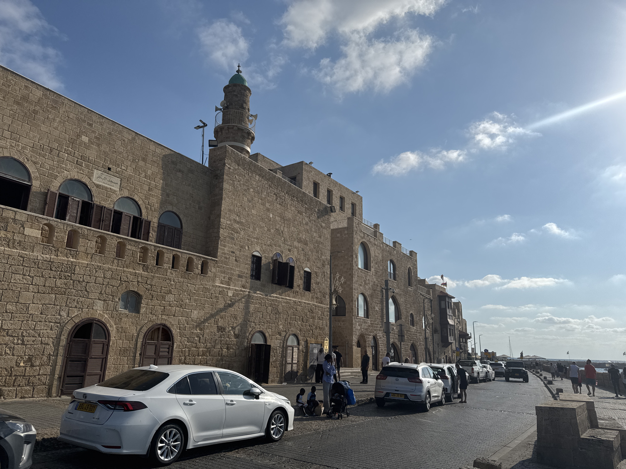 The image depicts a sunny day in the ancient city of Jaffa, a part of Tel Aviv, Israel, with a clear blue sky dotted with white clouds. The scene is dominated by a large, historic building constructed from rough-hewn, tan-colored stones. This building features arched windows, some with dark wooden shutters, and a prominent minaret with a green dome, suggesting it's a mosque.
In the foreground, a white sedan is parked on a cobblestone street. Several other vehicles, including SUVs and a jeep, are also parked along the street. People are visible walking along the waterfront promenade to the right, and a few individuals and children are gathered near the building on the left. A stroller is also present. The waterfront promenade appears to be paved with cobblestones and lined with low stone walls. In the background, the sea is visible, and a sailboat can be seen in the distance. The overall impression is one of a historical urban area with vibrant street life.