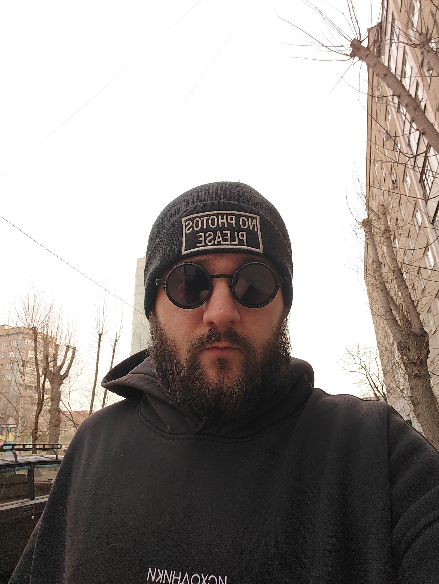 A man stands outdoors with buildings and trees in the background. He wears a black beanie with a rectangular patch that reads "NO PHOTOS PLEASE" in reverse, along with round, black sunglasses and a black hooded sweatshirt. He has a dark beard. The backdrop features multi-story buildings with bare trees suggesting it is not summer, and part of a black car is visible on the left. The location is Krasnoyarsk, Russia.
