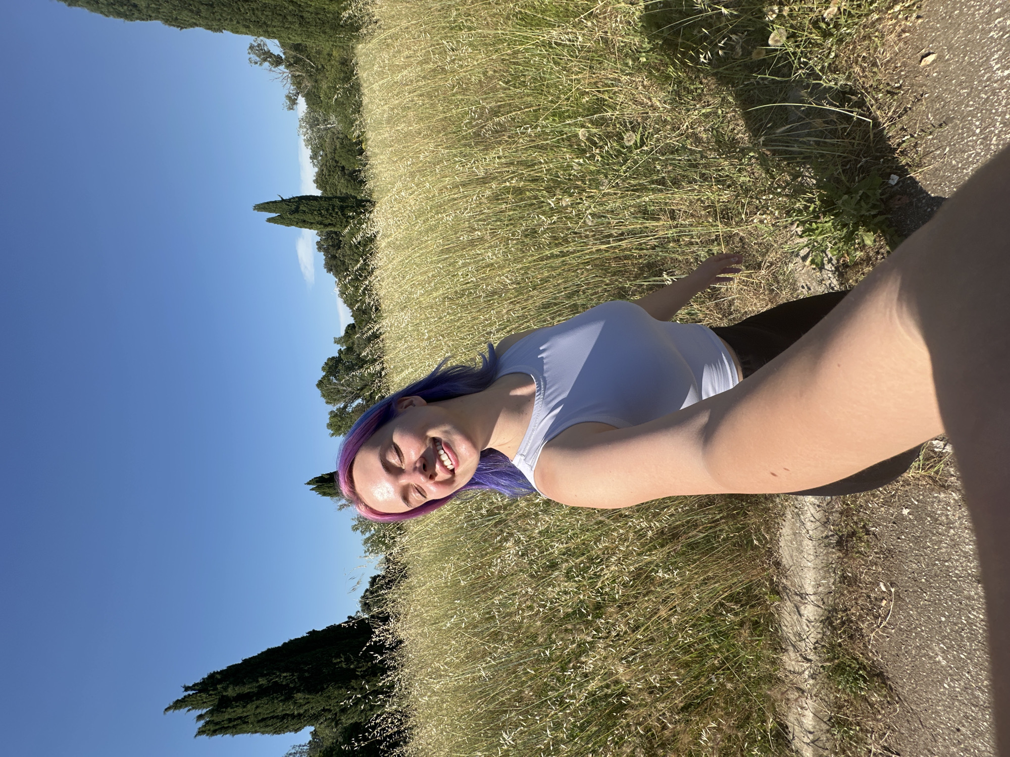 A young woman with dyed purple hair is taking a selfie in a grassy field in Caissargues, France. She is smiling and wearing a white top. The sky is blue and there are trees in the background. A paved surface is visible to the side.