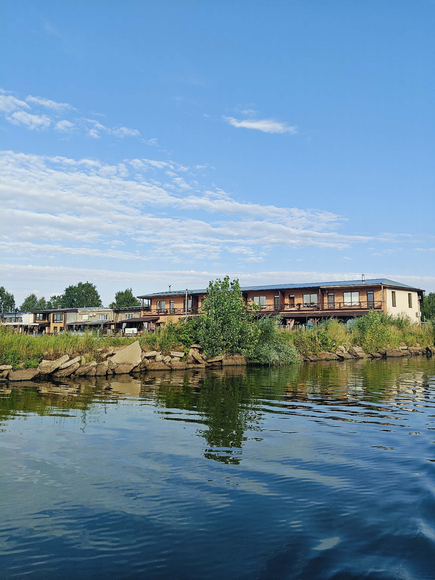 The image shows a row of modern, two-story houses situated along a waterfront in Chaikovsky, Russia. The houses are made of brick and have balconies and large windows. They are surrounded by green vegetation, including trees and shrubs. In the foreground, there is a body of water with visible ripples. The sky above is blue with scattered white clouds. The scene appears to be a residential area, possibly a resort or a private development, located on the banks of a river or lake.