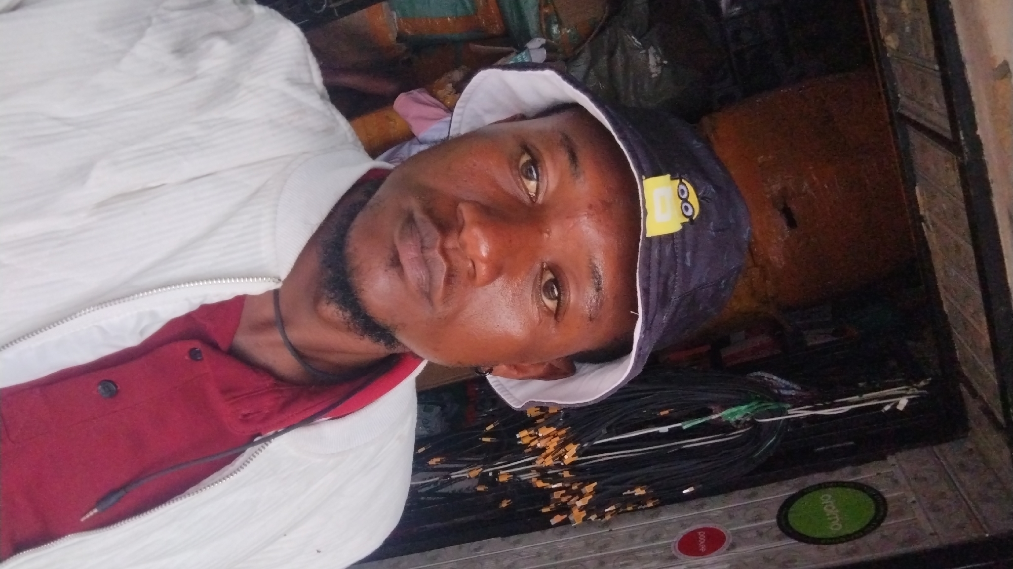 The image shows a person wearing a white jacket, red shirt, and a dark-colored hat with a Minion cartoon character on it. The person has a beard. The background includes items possibly stored in a room or storage area, with various objects like boxes, cables with connectors, and branded stickers on the floor. The location, Jos, Nigeria, is not explicitly identifiable from the image; the environment suggests an indoor storage area. There is no immediate information on specific actions or interactions.