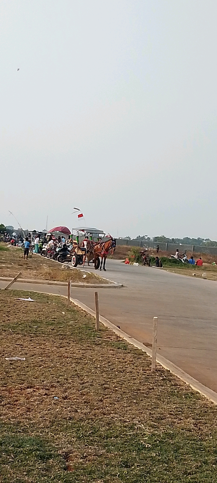 The image depicts a scene in Satriamekar, Indonesia, on a bright, overcast day. In the foreground, dry, brown grass lines a paved road. Wooden posts are visible along the roadside. In the middle ground, a horse-drawn carriage with a white roof and red accents, pulled by two brown horses, is moving along the road. Several motorcycles and people are gathered around a canopy with a red umbrella, likely a marketplace or event. A red and white flag is visible on a pole behind the carriage. In the background, more people are seated or standing around a fence, with some individuals on motorcycles. Trees and vegetation are present behind the fence, and the sky is a pale, hazy white. A small, dark object, possibly a kite or bird, is visible in the upper left corner of the sky.