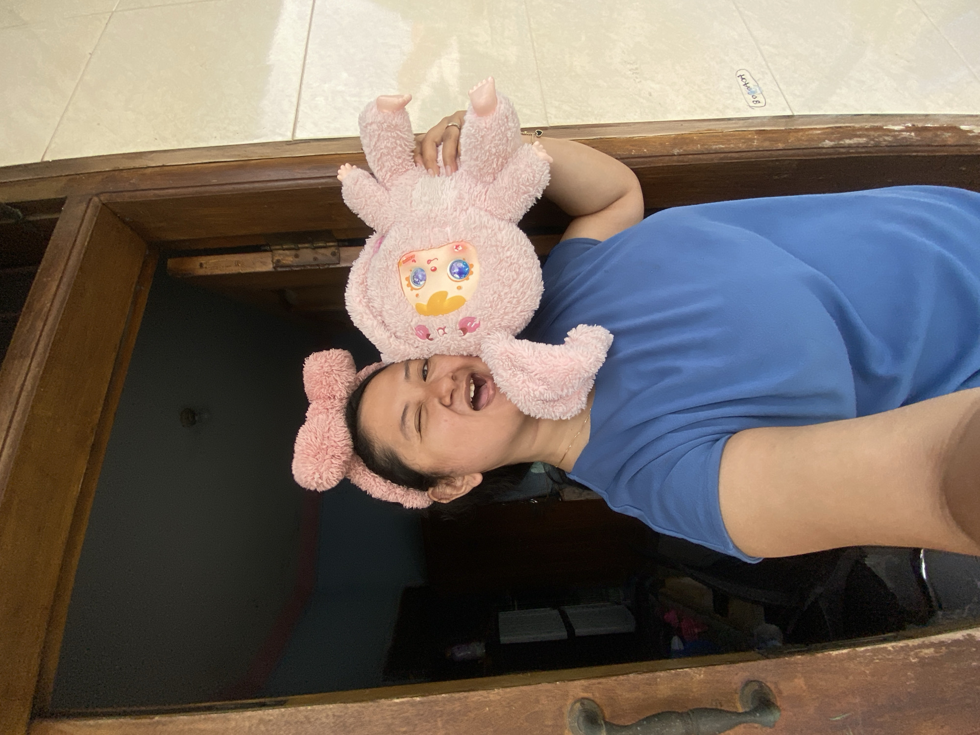 A medium-shot, rotated image depicts a person, likely female, lying down indoors, looking up towards the camera. She wears a blue short-sleeved top and a pink fuzzy headband with large ears. Her mouth is open in a joyful or laughing expression, and her eyes are squinted with amusement. Resting on her forehead and hair is a plush, pink, rabbit-like toy featuring a yellow face with large, blue, sparkly eyes and small pink cheeks. A gold chain is visible around her neck, and a ring is on her finger, which is partially seen holding the plush toy.
The person is positioned next to a dark, worn wooden frame, which runs horizontally across the lower and upper thirds of the image. Above this wooden frame, a light-colored tiled floor is visible. On this tiled surface, near the person's right shoulder, there is text written in an oval shape, which reads "Bongovoz". Below the wooden frame, the background is a dark, indistinct interior space. In the lower right corner, a portion of a dark metal handle is visible on the wooden frame. The lighting suggests an indoor setting during the daytime or with artificial light. The overall mood is playful and cheerful.