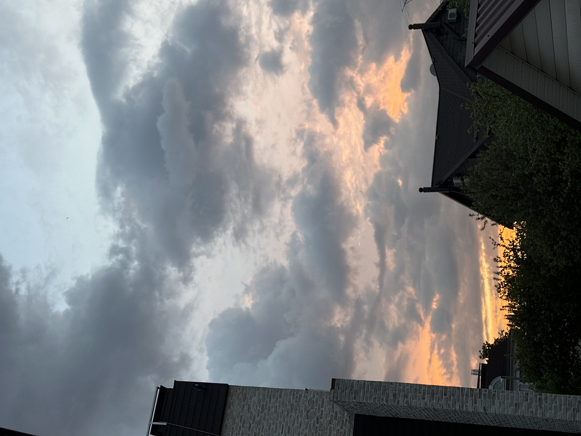 The image shows a sky with dark and light clouds at sunset. The sky exhibits a range of colors from light grey to orange, indicating the setting sun. The view appears to be from a residential area in Nizhny Novgorod, Russia. The built environment includes buildings with dark roofs, brick walls, and green vegetation. The orientation is vertical, suggesting a photograph taken from a lower perspective.