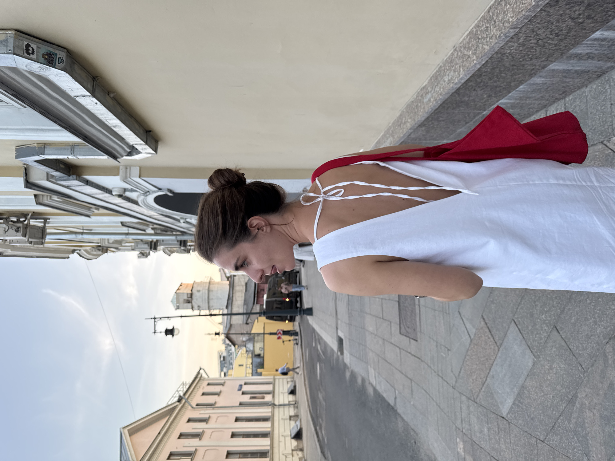 A medium shot of a woman on a sidewalk in Moscow, Russia. She has brown hair and is wearing a white dress with spaghetti straps and a red bag over her shoulder. She is looking down and appears to be walking. The sidewalk is made of grey paving stones. Buildings are visible in the background, including a light yellow building with windows and a taller building with a tower. There is a streetlamp on the sidewalk. The sky is slightly cloudy.