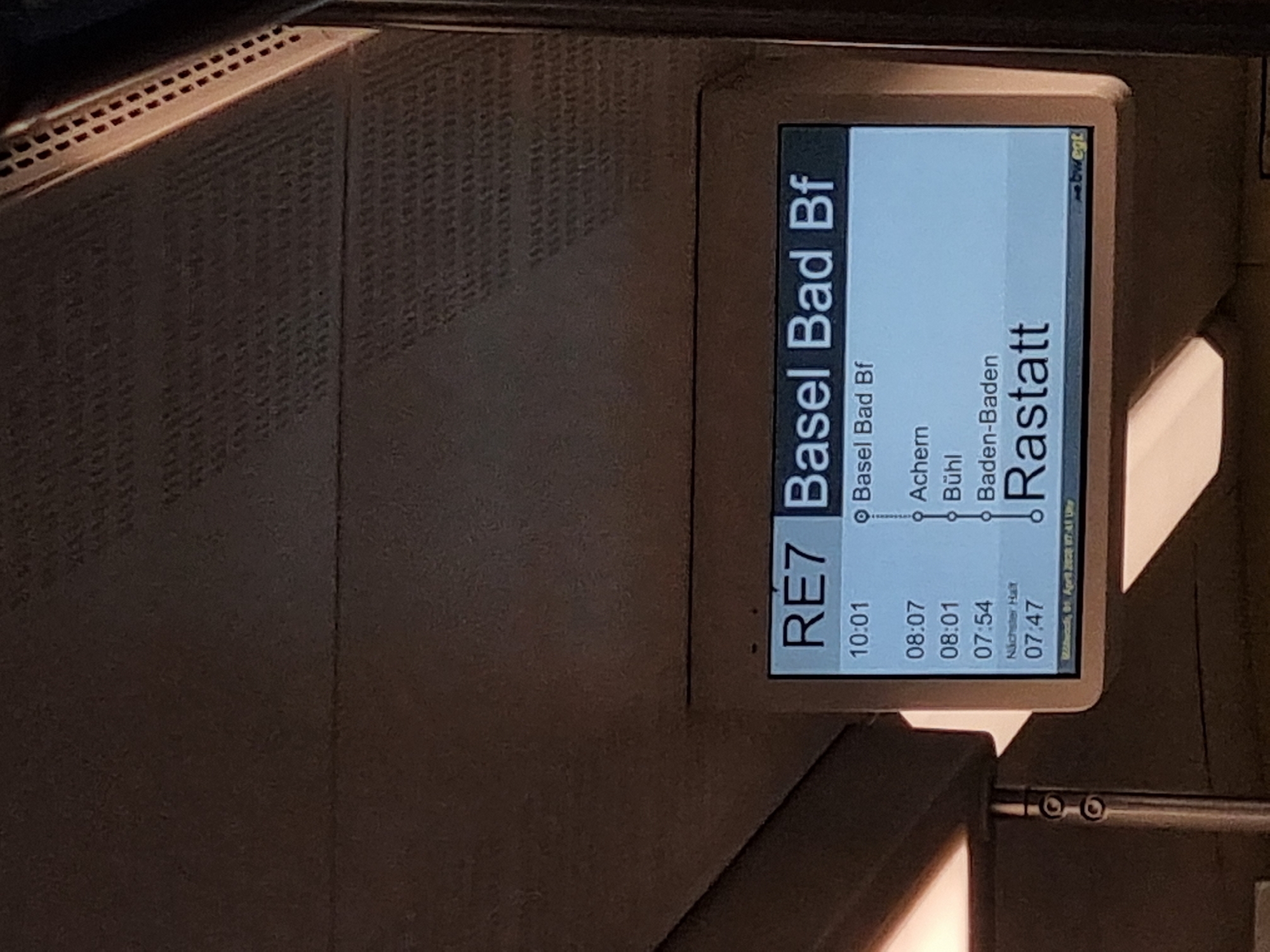A digital display shows a train schedule with the heading "RE7 Basel Bad Bf." The display indicates the train's route, showing stops at Basel Bad Bf, Achern, Bühl, Baden-Baden, and Rastatt. Scheduled departure times are listed next to each stop. The display notes the next stop is at Rastatt, scheduled for 07:47. The date shown at the bottom of the display reads "Wednesday, 01 April 2028 17:41 Uhr". The location is not stated.