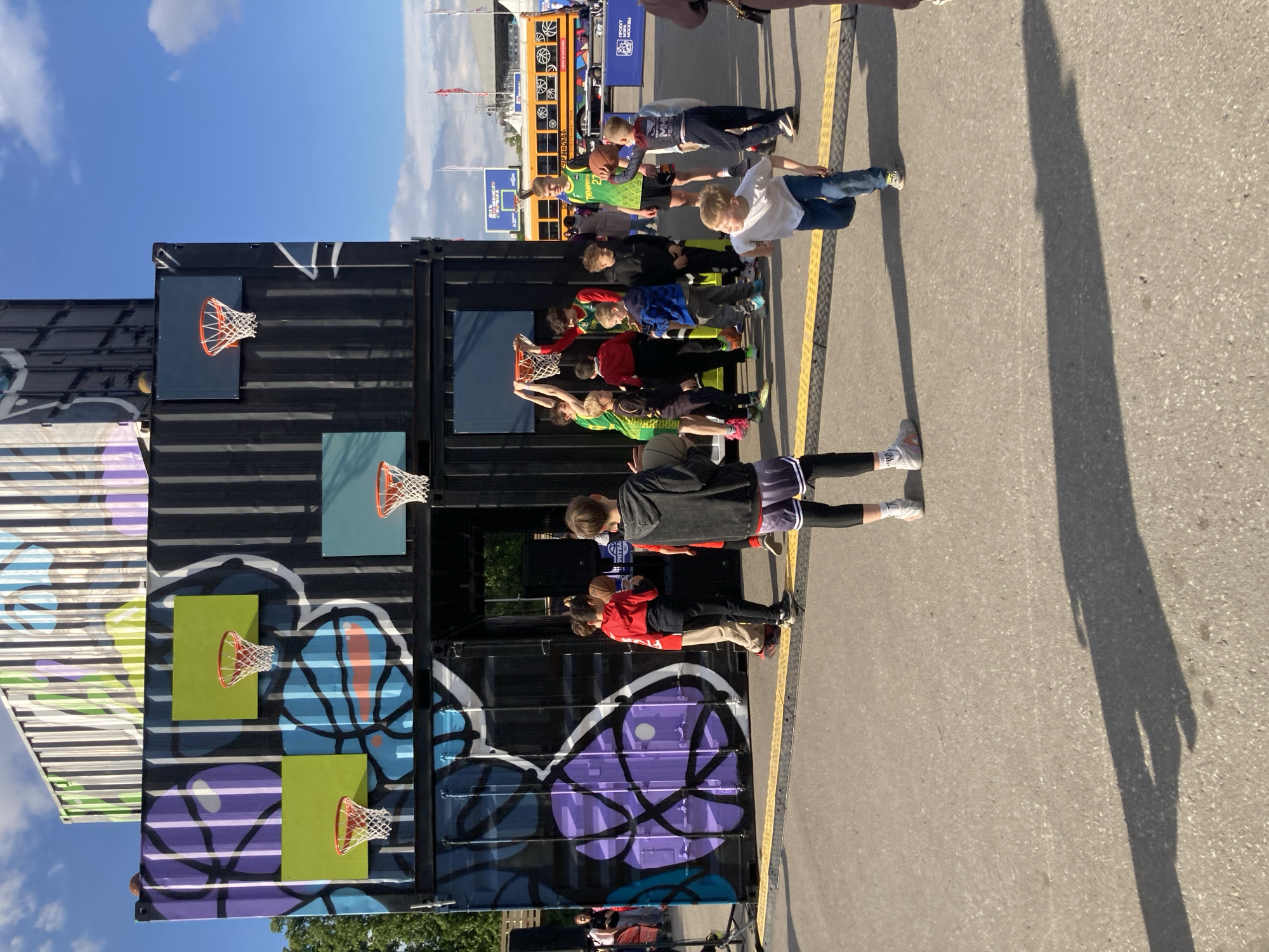Here's a description of the image:
The photograph shows a group of children standing in a line, presumably waiting their turn, at a mobile basketball setup. The basketball setup is made of shipping containers with graffiti art and multiple basketball hoops attached. The scene is outdoors, on an asphalt surface marked with a yellow line. In the background, a blue school bus and what appears to be a large event venue can be seen. Based on the information on the bus, the location is Moscow, Russia. The children are wearing a variety of casual clothing, including sports jerseys, T-shirts, and jeans. Some children are holding basketballs.