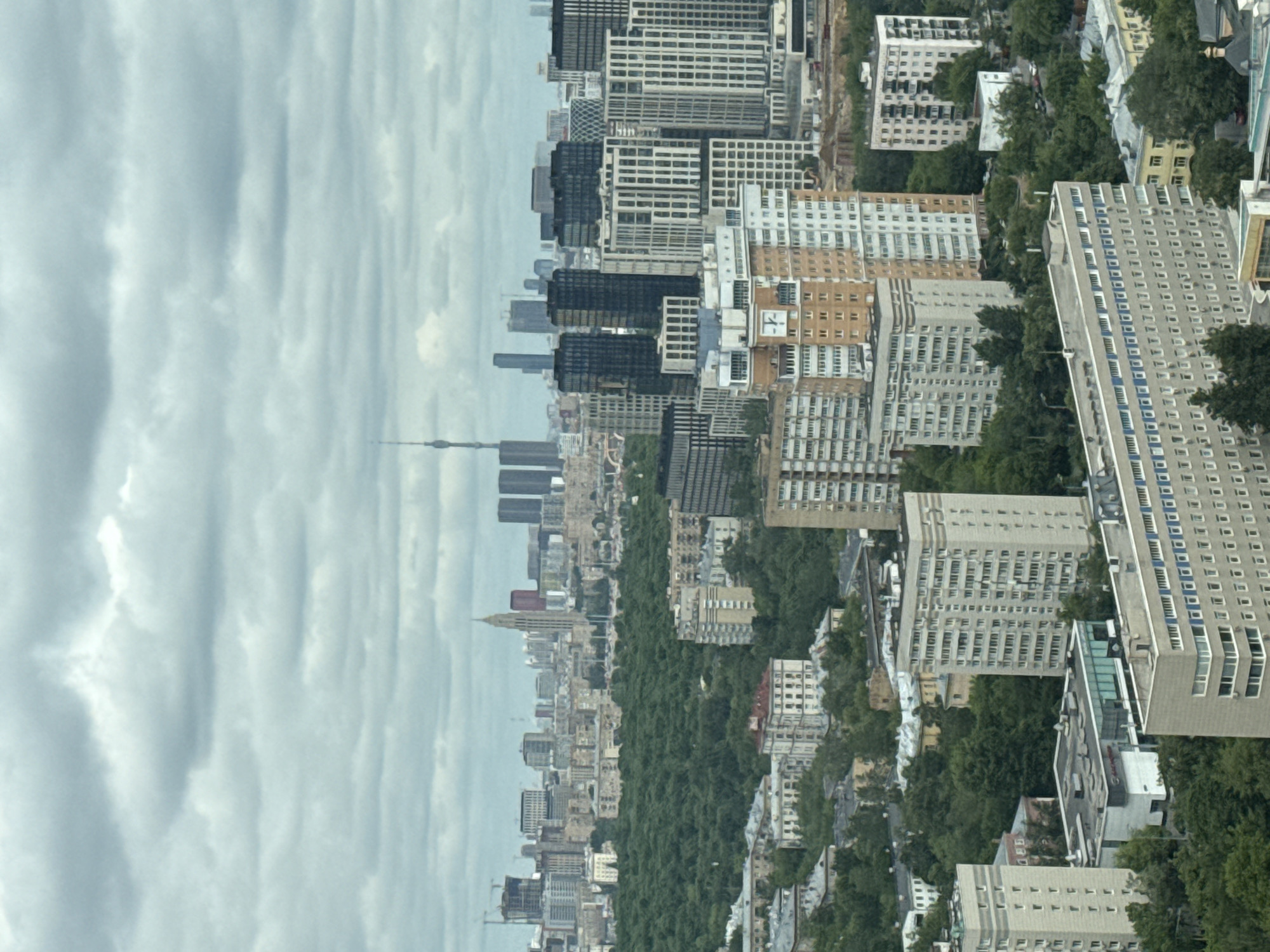 An aerial, high-angle view captures a sprawling cityscape under an overcast sky. The scene is dominated by a diverse collection of buildings, ranging from modern dark-facade high-rises to lighter-colored, multi-story residential and commercial structures. In the far distance on the left, a tall, slender tower, identifiable as the Ostankino Tower given the location context of Khimki, Russia, stands prominently against the cloudy horizon.
Extensive stretches of dense green tree canopy are interspersed throughout the urban fabric, suggesting large parks, boulevards, or forested areas that weave through the city's developments. In the middle ground and foreground, various buildings display different architectural styles, some with intricate window patterns and balconies. One notable detail on a beige building in the mid-right of the frame is a clock face with Roman numerals, although the exact time it displays is not clearly discernible.
The weather appears overcast with soft, diffuse light indicating daytime. No specific human activity or people are visible due to the high vantage point. There is no readable text present on any of the buildings or signs in the image. The overall impression is a vibrant, well-developed urban environment harmoniously integrated with significant green spaces.