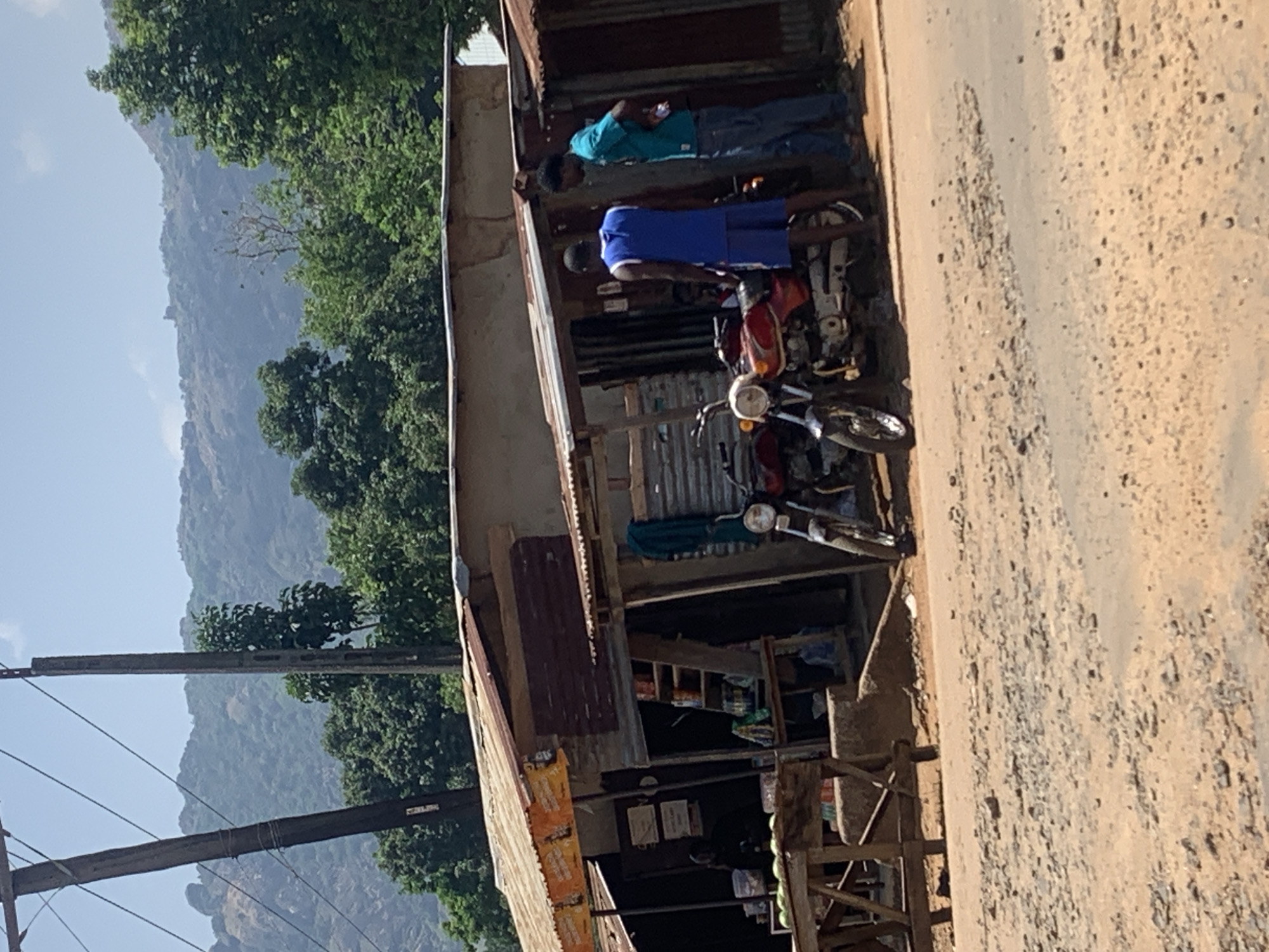 The image shows a roadside scene in Kukum, Nigeria. Two men stand in front of a corrugated metal structure that appears to be a small shop. One man wears a blue shirt, and the other wears a turquoise shirt. Two motorcycles are parked nearby. The road is unpaved and rocky. In the background, trees and a mountain are visible. A power pole is also visible on the left side of the frame.
