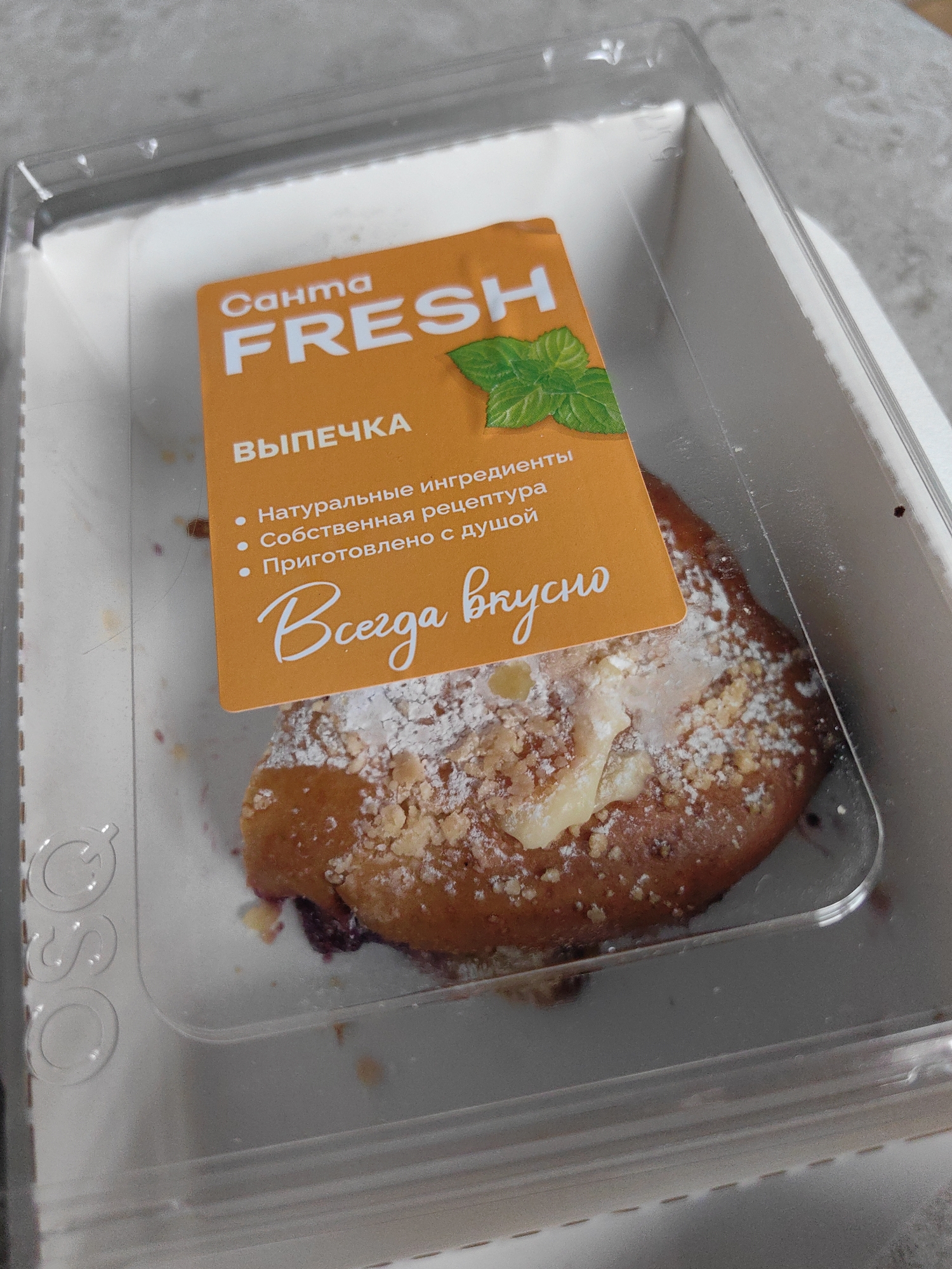 A close-up, high-angle shot shows a baked pastry, likely a bun, dusted with powdered sugar and some crushed nuts, sitting inside a clear plastic container. The pastry is on a white cardboard base. A bright orange label is partially obscuring the pastry. The label has text in Russian: "Санmа FRESH" (Sante FRESH) and "ВЫПЕЧКА" (Baking). Below that, bullet points read "Натуральные ингредиенты" (Natural ingredients), "Собственная рецептура" (Proprietary recipe), and "Приготовлено с душой" (Made with soul). At the bottom of the label, in a cursive font, is the phrase "Всегда вкусно" (Always delicious). A small illustration of mint leaves is also visible on the label. The clear plastic lid of the container is visible, and the overall environment suggests an indoor setting, possibly a store or home kitchen. The image does not contain any elements that would indicate a specific location in the city of Fanipal, Belarus.