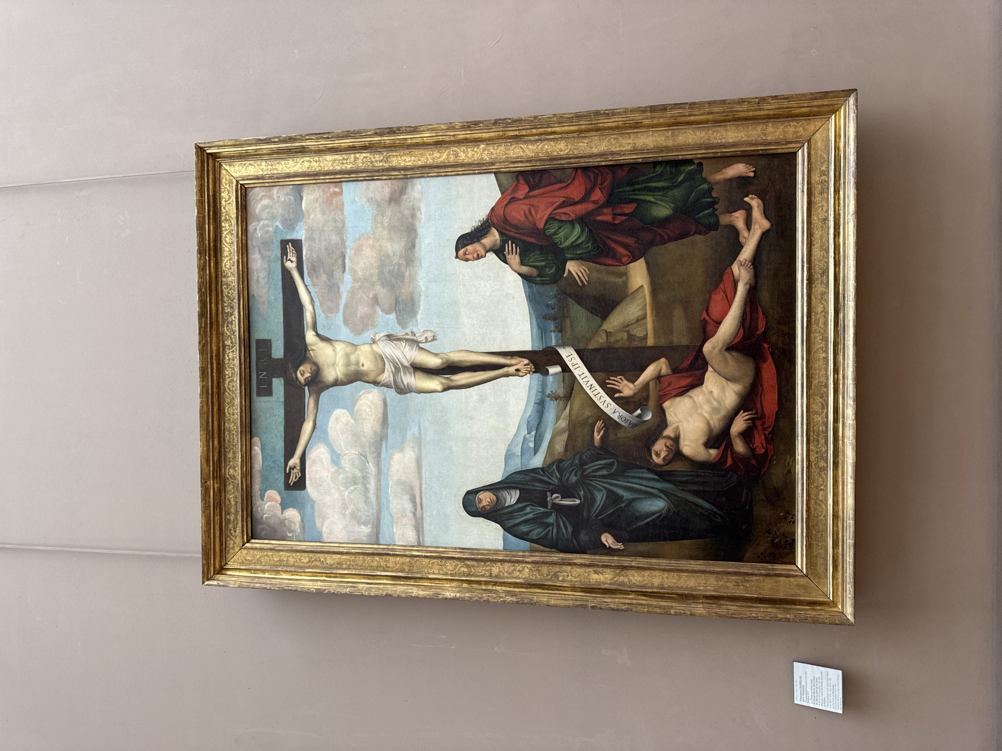 The image shows a framed painting hanging on a neutral-colored wall. The painting depicts the crucifixion of Jesus Christ. Jesus is shown on a cross in the center. At the foot of the cross is a mourning figure dressed in dark clothing. Another figure is partially supporting Jesus's body. Above them, a figure in red and green robes is partially visible. The painting has a detailed, ornate gold frame. A small informational card is attached to the wall below the painting. The painting is located in the Louvre Museum in Paris, France.