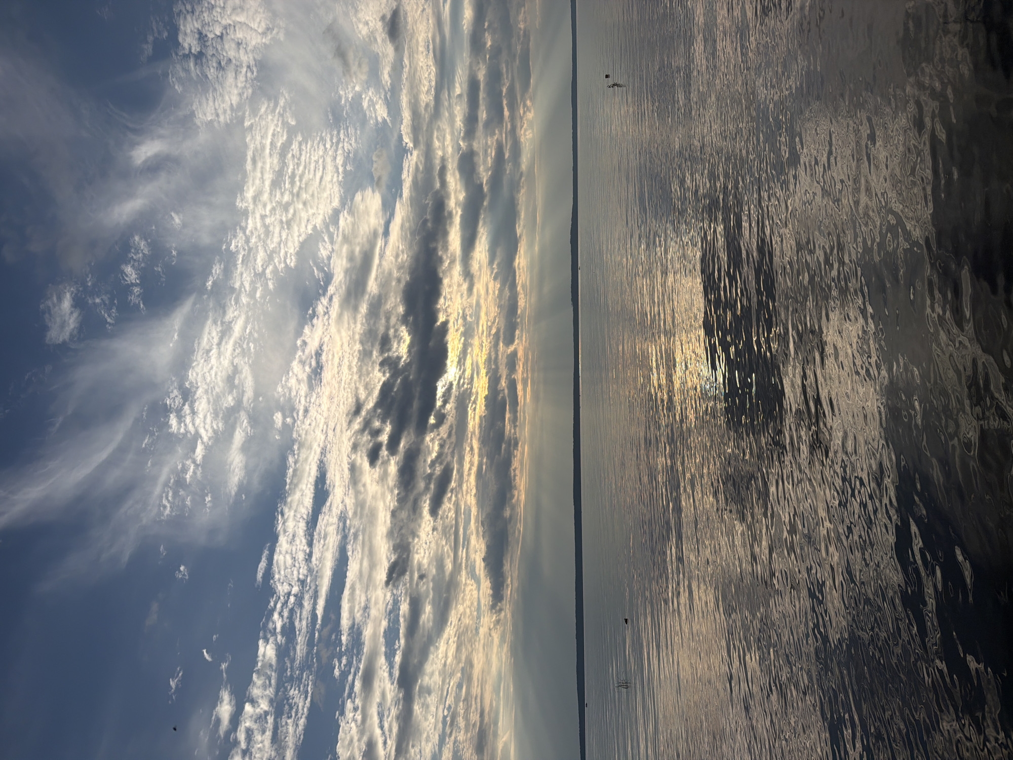 The image depicts a serene, wide-angle view of a body of water, likely a lake or large river, under a dramatic sky. The sun is partially obscured by clouds, casting visible sunbeams through breaks in the cloud cover, creating a dramatic lighting effect on the water's surface. The sky is a mix of blue and white, with wispy clouds that suggest a gentle breeze.
In the distance, a faint, dark line marks the horizon, indicating land. The water is calm, with ripples reflecting the sky and sunlight. There are a few small, indistinct objects on the water's surface, possibly buoys or small vessels, and a single, dark, vertical element, possibly a branch or a small structure, in the mid-left of the frame.
The overall impression is one of natural beauty and tranquility, possibly captured during sunrise or sunset due to the quality of the light. The location is specified as Stepanovo, Russia.