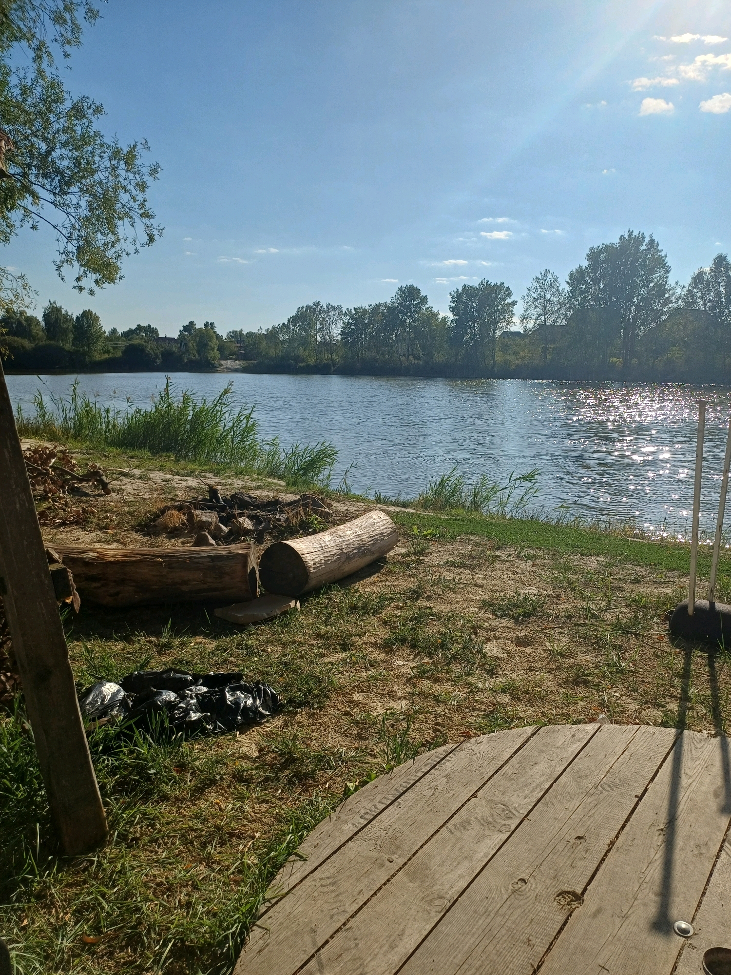 The image depicts a serene outdoor scene by a body of water, likely a river or lake, situated in Sulymivka, Ukraine. The foreground is dominated by a wooden structure and grassy terrain, with two large logs lying on the ground, one halved lengthwise. A campfire pit with stones and charred wood is visible between the logs. Black trash bags are present near the campfire. The middle ground features a sandy bank with tall green reeds bordering the water. The water itself is calm, with sunlight reflecting off its surface, creating a shimmering effect. The background showcases a dense line of trees and vegetation, with a few houses partially visible amongst them. The sky is clear blue with scattered white clouds, and sunlight streams down, casting shadows. The overall impression is one of a peaceful natural setting, possibly a recreational area.