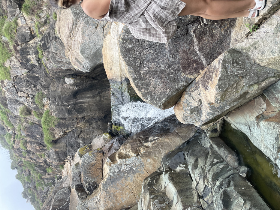 The image displays a natural waterfall within a rugged, rocky environment identified as being in Ninh Hòa, Vietnam. In the right foreground, a single individual is partially visible from the back, facing towards the waterfall. The person wears a light-colored, open plaid shirt, revealing a bare right shoulder and a beaded necklace, along with white open-toed sandals. The landscape features numerous large, angular rock formations, varying in color from dark grey to light tan, with visible patches of green moss and sparse vegetation on the upper slopes and crevices. A torrent of white water cascades downwards in the center-left, flowing into small, dark green pools at various levels among the rocks. A white rope or cable is visible on the upper left side, secured to a rock.
