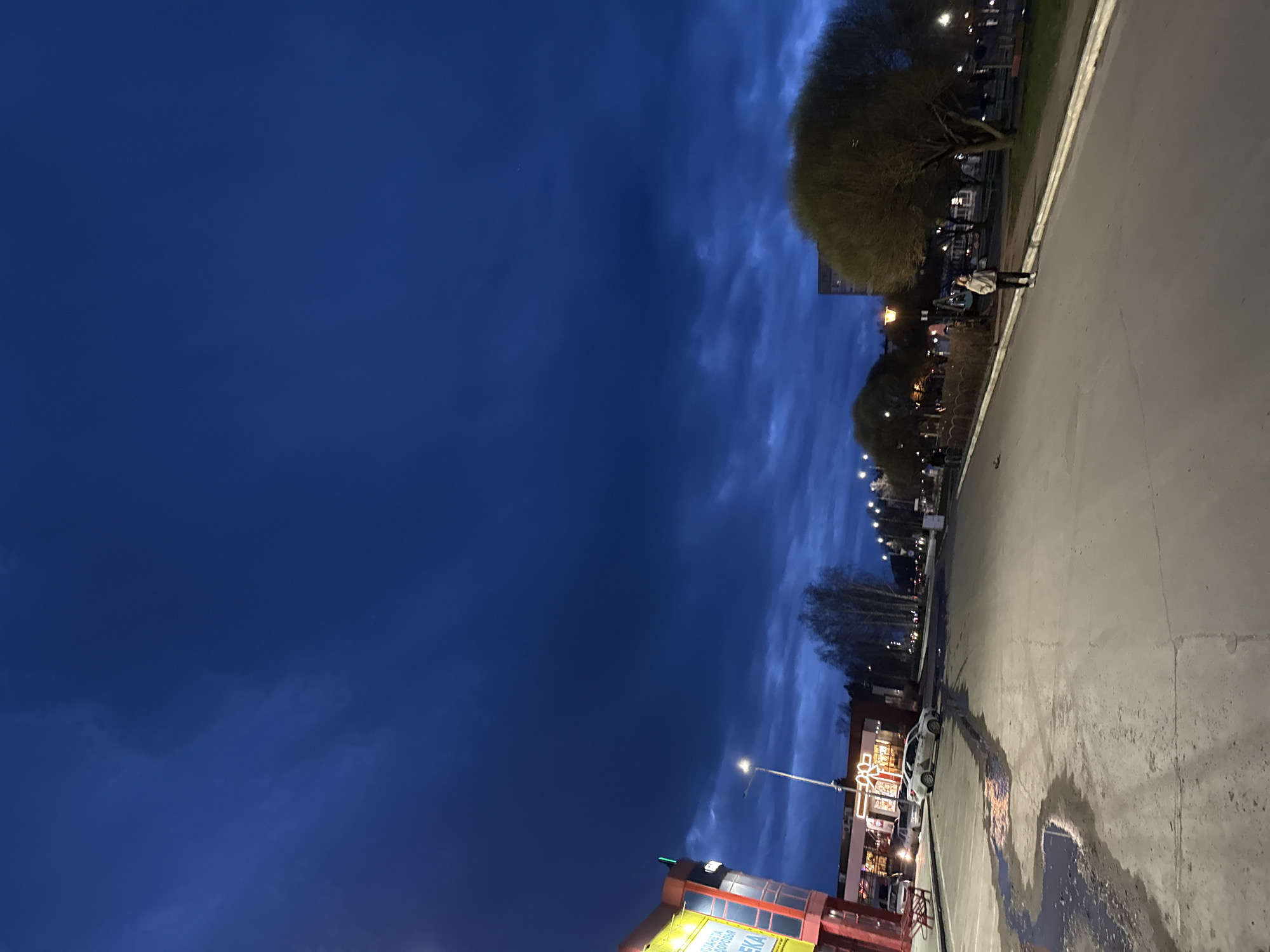 The image depicts an urban scene at dusk or night in an unspecified city. The sky is a deep blue with streaky clouds. Buildings and trees line the right side of the image along a concrete sidewalk or parking area, which has patches of wetness. Streetlights illuminate the area. There is a person standing in the distance, as well as parked vehicles visible. The city is Unknown.