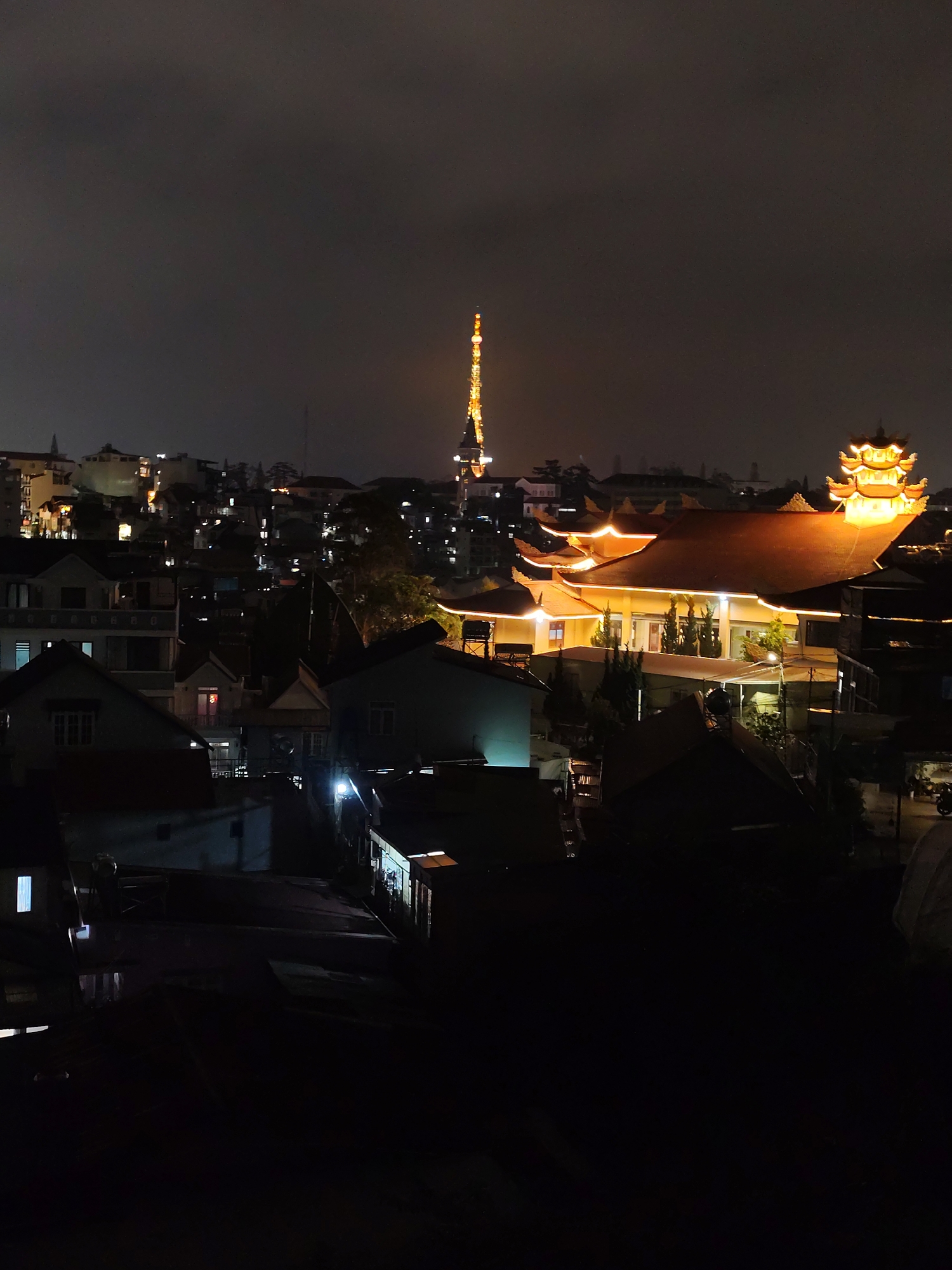 At night, a residential area of Da Lat, Vietnam, is illuminated. The built environment comprises many low-rise buildings. The city's telecommunications tower, lit up in gold, rises above the houses. A traditional temple stands out, glowing gold against the darkness. The lighting suggests some activity, but the area mostly appears serene and quiet.