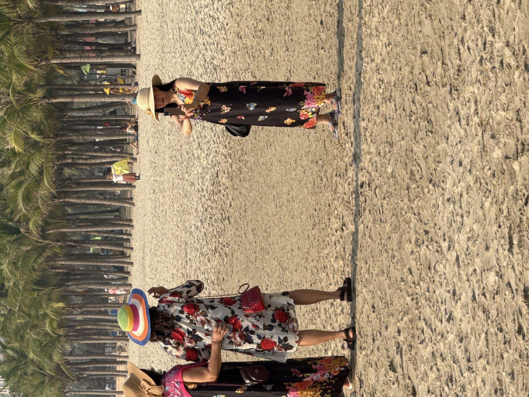The scene shows a sandy beach lined with palm trees, likely in Đà Nẵng, Vietnam. Three women are seen on the beach. The woman in the center wears a short white floral dress and a colorful straw hat. One woman is taking a photo with her cell phone wearing a black floral dress and tan straw hat. The third woman is partially visible, wearing a dark dress with a pink pattern.
