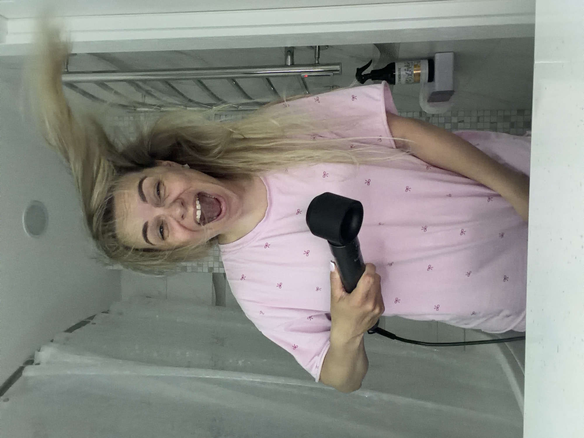 A fair-skinned woman is holding a black hair dryer in her right hand while standing in a bathroom. She is tilting her head towards her left shoulder, and her mouth is open in an apparent yell. Most of her long blonde hair is being flung upwards and to the left. She is wearing a pink t-shirt that has a pattern of tiny bows on it. Behind her, there is a light grey wall. A metal rack and a bottle of hair product are mounted on the wall. A shower curtain is visible in the foreground. The location of the photograph is not identifiable as being in Votkinsk, Russia, or any other specific location.