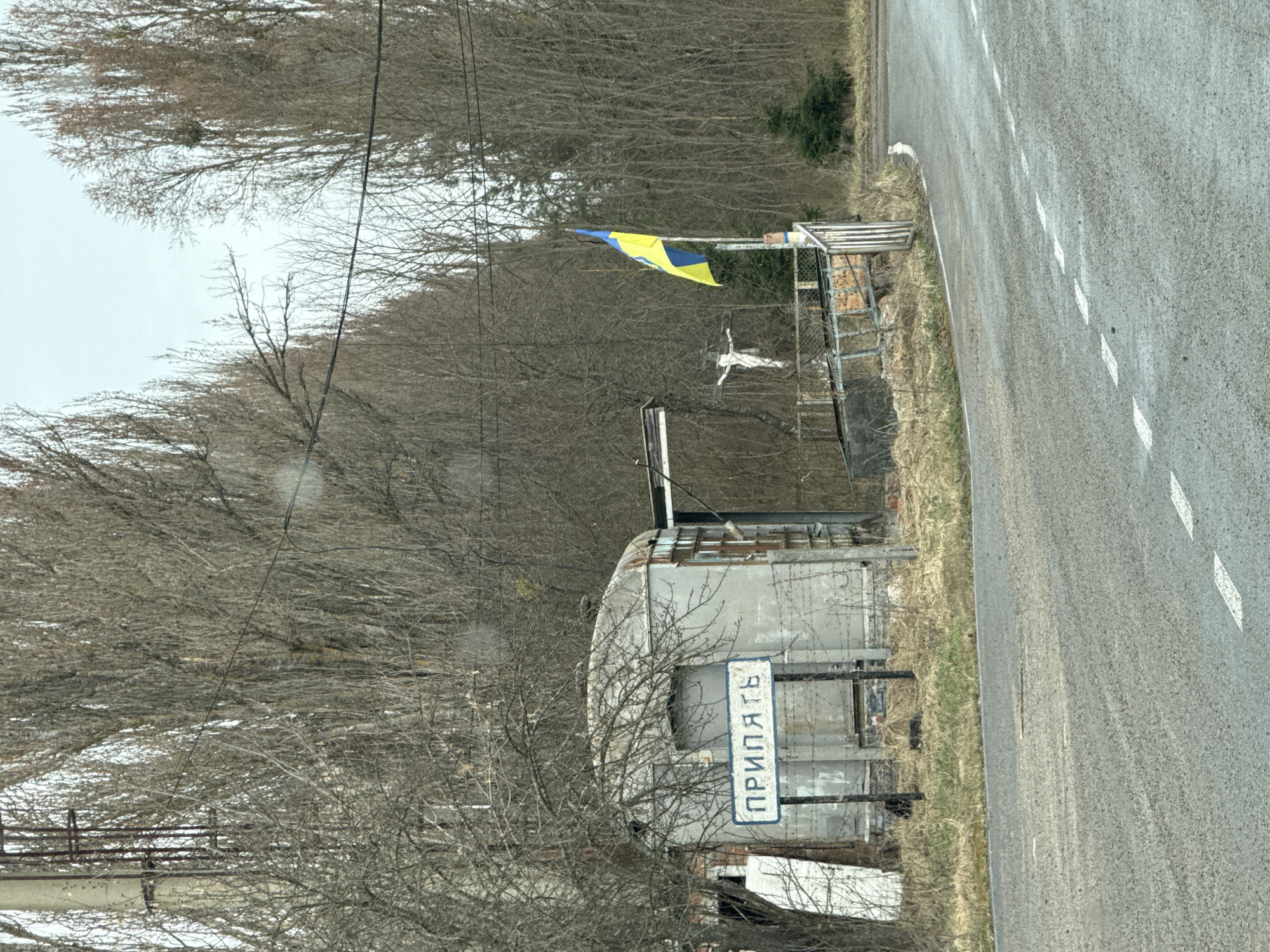 The image shows the city entrance sign to Prypiat, Ukraine, positioned on the side of a road. The sign, bearing the city's name in Cyrillic, is mounted on a dilapidated structure. A Ukrainian flag is visible. The environment includes bare trees and a gray road. The scene is desolate and suggests neglect or abandonment.