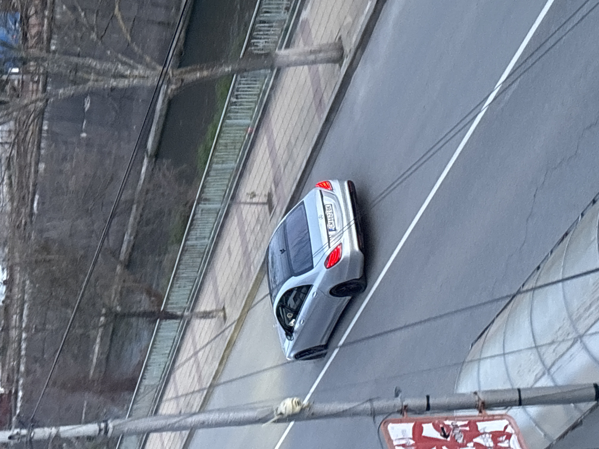 The image depicts a silver Mercedes-Benz car driving on a road alongside a river or canal in Zlatograd, Bulgaria. The car, partially angled, has a license plate that reads "CM 6622 BK." A white lane marker is visible on the road. On the left side, a railing runs along the river/canal, with bare trees and power lines visible in the background. In the foreground, there are parts of what appears to be a street sign or advertisement.