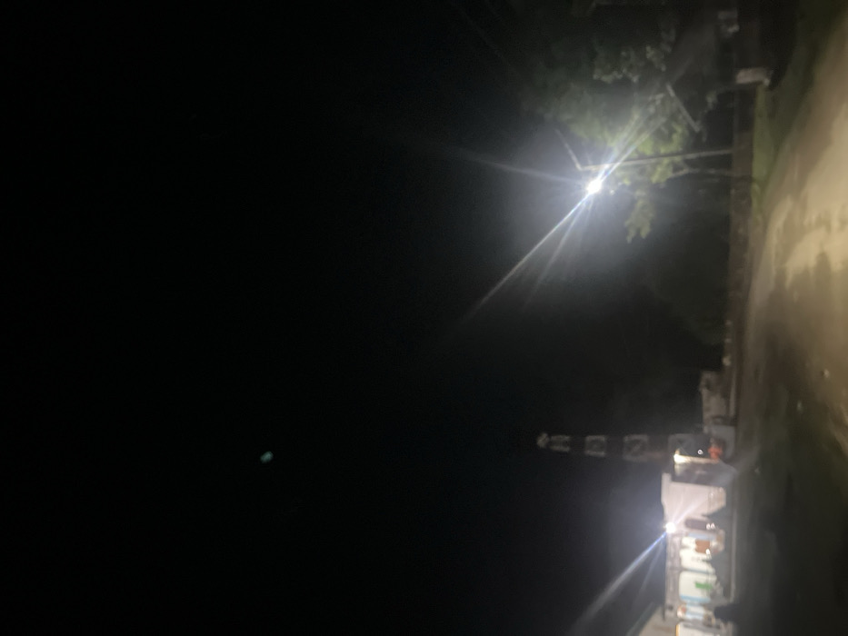 A night scene displays a stark contrast between deep black darkness and bright artificial light. A tall, latticed tower stands illuminated amidst the blackness. A streetlamp shines intensely near green foliage, casting light rays across the dark sky. A wet-looking road runs to the side. A building, possibly a shop, is lit from within, with bright light spilling out.