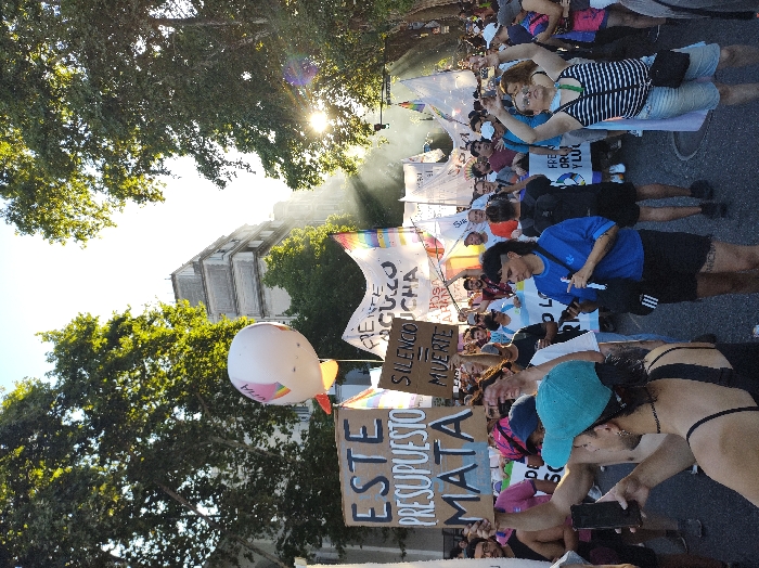 A crowd of people marching on a sunny day. Some people are lying on the ground. Many are holding signs, including one that says "Este Presupuesto Mata" (This Budget Kills) and "Silencio = Muerte" (Silence = Death). There are rainbow flags, banners, and a large white balloon. Trees line the street, and a tall building is visible in the background. The scene appears to be a protest or demonstration.