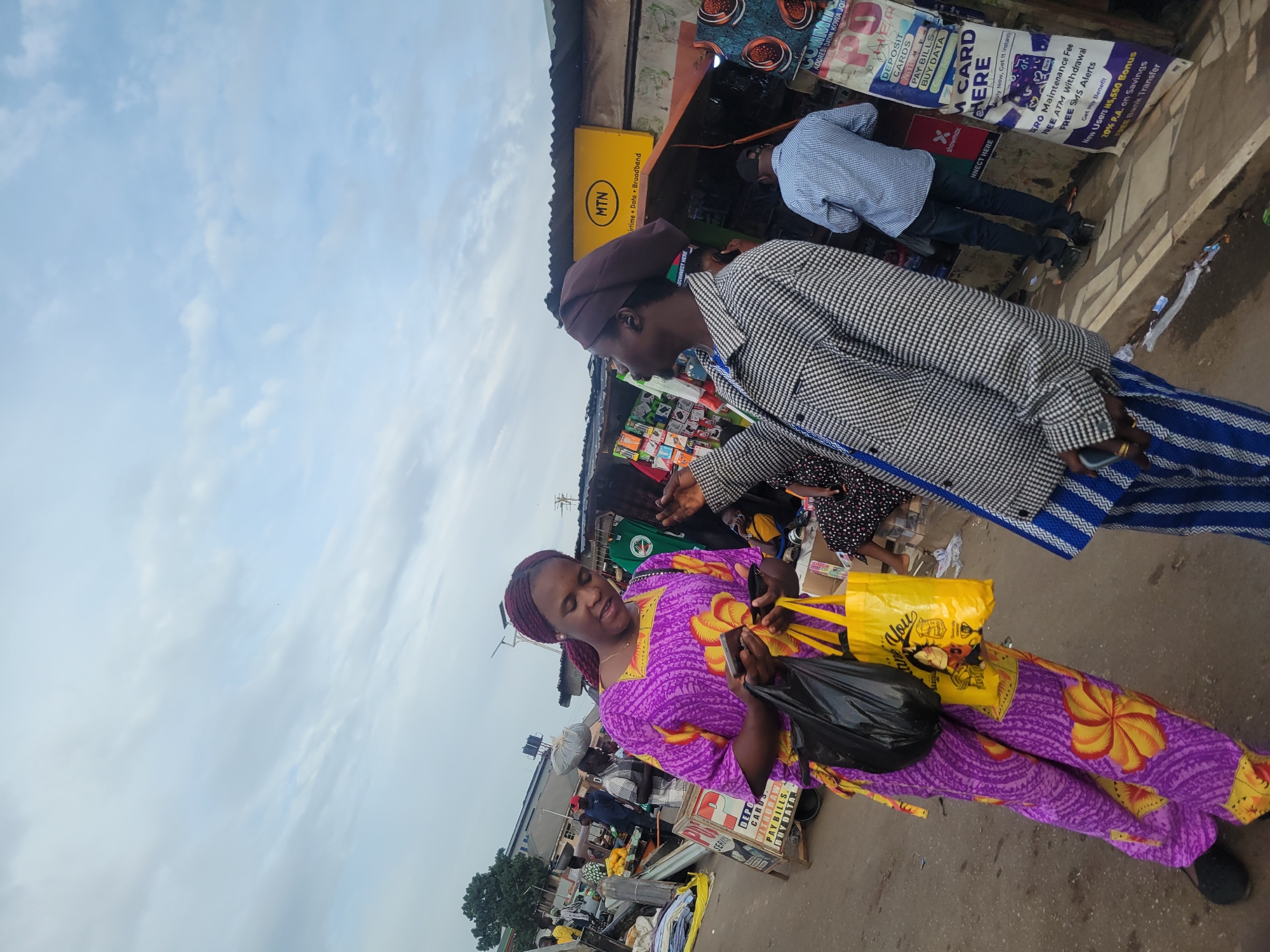 The image depicts a bustling street market scene in Jos, Nigeria, under an overcast sky. In the foreground, a woman wearing a vibrant purple and yellow patterned dress holds a black plastic bag and a yellow shopping bag, which has "Thank You" printed on it. She appears to be focused on a mobile phone in her hand. Facing her is another person, dressed in a checkered jacket and a brown beret-style hat, suggesting an interaction or transaction between them.
The background reveals an active market environment with several makeshift stalls and vendors. A prominent yellow sign for "MTN Airtime + Data + Broadband" is visible, indicating telecommunication services. Numerous other signs advertise financial and mobile money services, with text such as "DEPOSIT CARDS," "WITHDRAW," "PAY BILLS," "BUY DATA," "M CARD HERE," "ZERO Maintenance Fee," "FREE ATM Withdrawal," "FREE SMS Alerts," "New Users N5,550 Bonus," "20% P.A. on Savings," and "FREE Bank Transfer." One sign also includes "showmax" and partially visible text "COMMUNITY ADDRESS DADIN KOWA JOS," confirming the location. Other market goods, including blue and white striped mats, are arranged for sale. The scene conveys a typical busy daytime atmosphere of an informal street market.