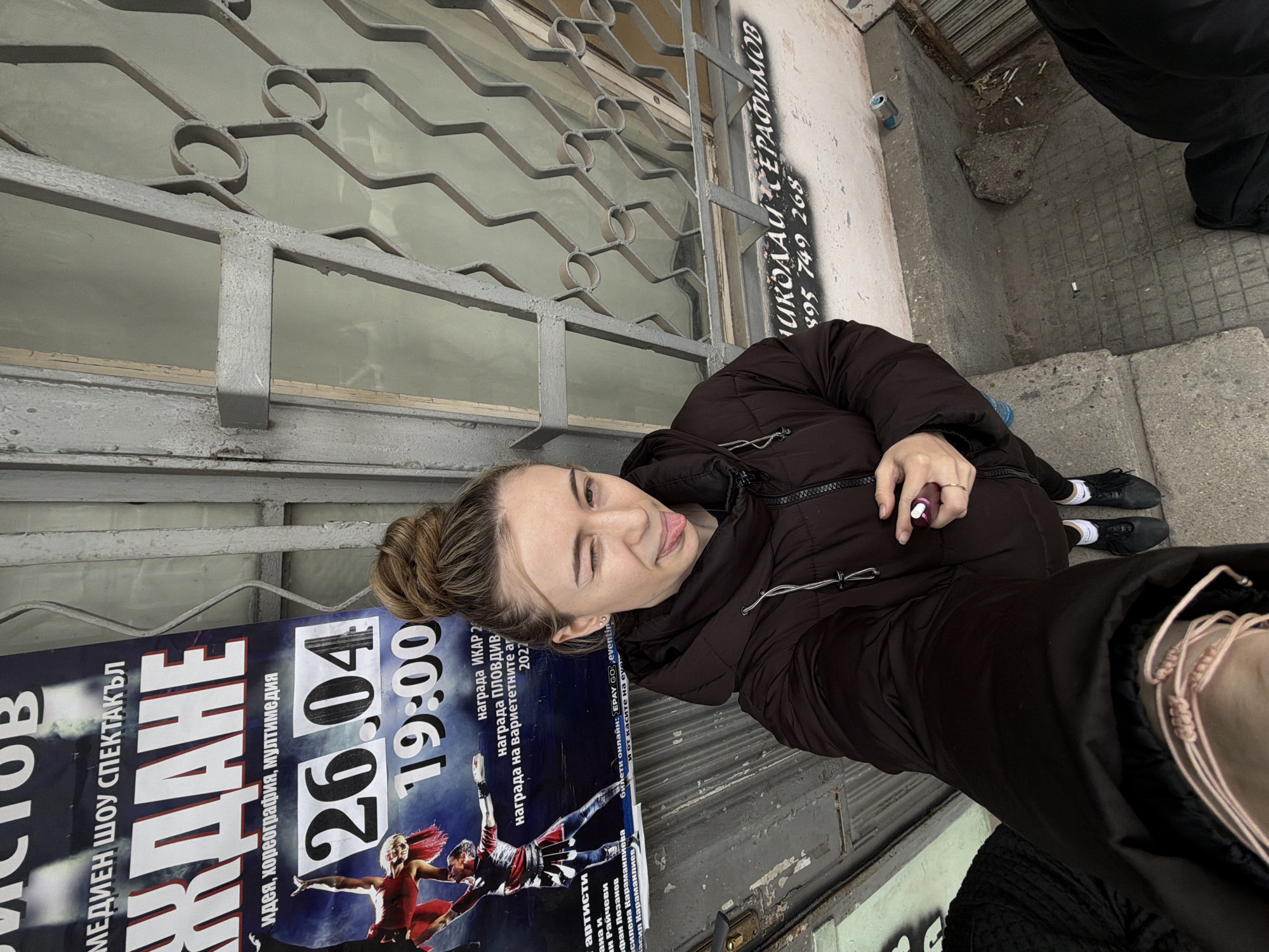 A woman stands beside a poster advertising a performance, likely a show or event, in Plovdiv, Bulgaria, judging by the Cyrillic text. The poster features the date "26.04" and time "19:00," and the woman appears to be posing for a photo or selfie near it. The poster advertises "ЖДАНЕ," which translates to "Waiting" in English. The environment appears to be an urban setting with a window protected by metal bars. The scene also shows a mobile number written on the wall along the poster.