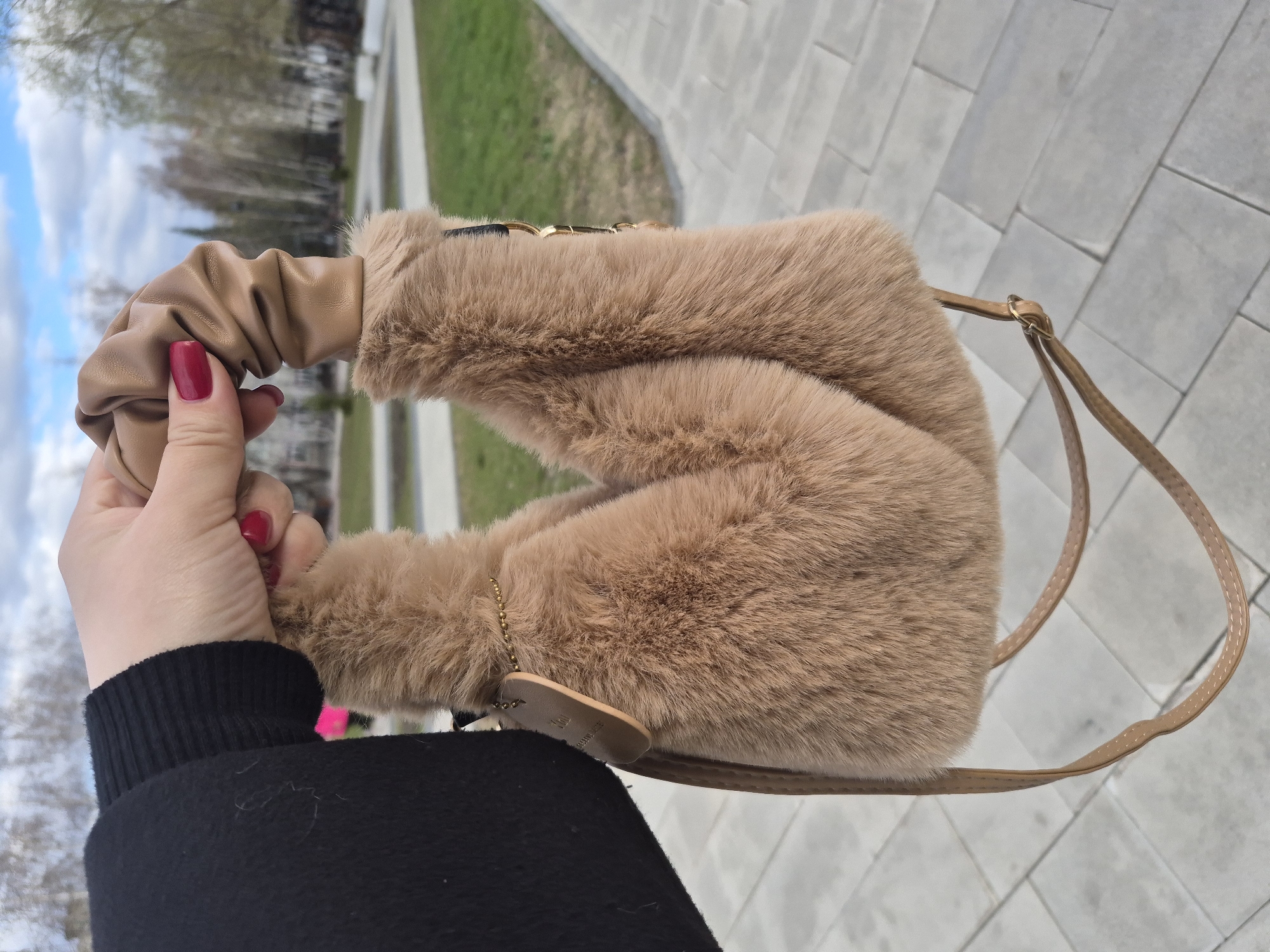 A person wearing a black long-sleeved top holds a beige faux-fur handbag by its beige faux-leather handle, with red painted fingernails visible on the hand. The bag has a beige faux-leather shoulder strap attached with metal loops. A small, unreadable leather tag hangs from a thin gold chain. The setting is an outdoor urban environment, specifically Tobolsk, Russia, with a gray brick walkway beneath the bag. Background elements include greenery, trees, and a cloudy sky.