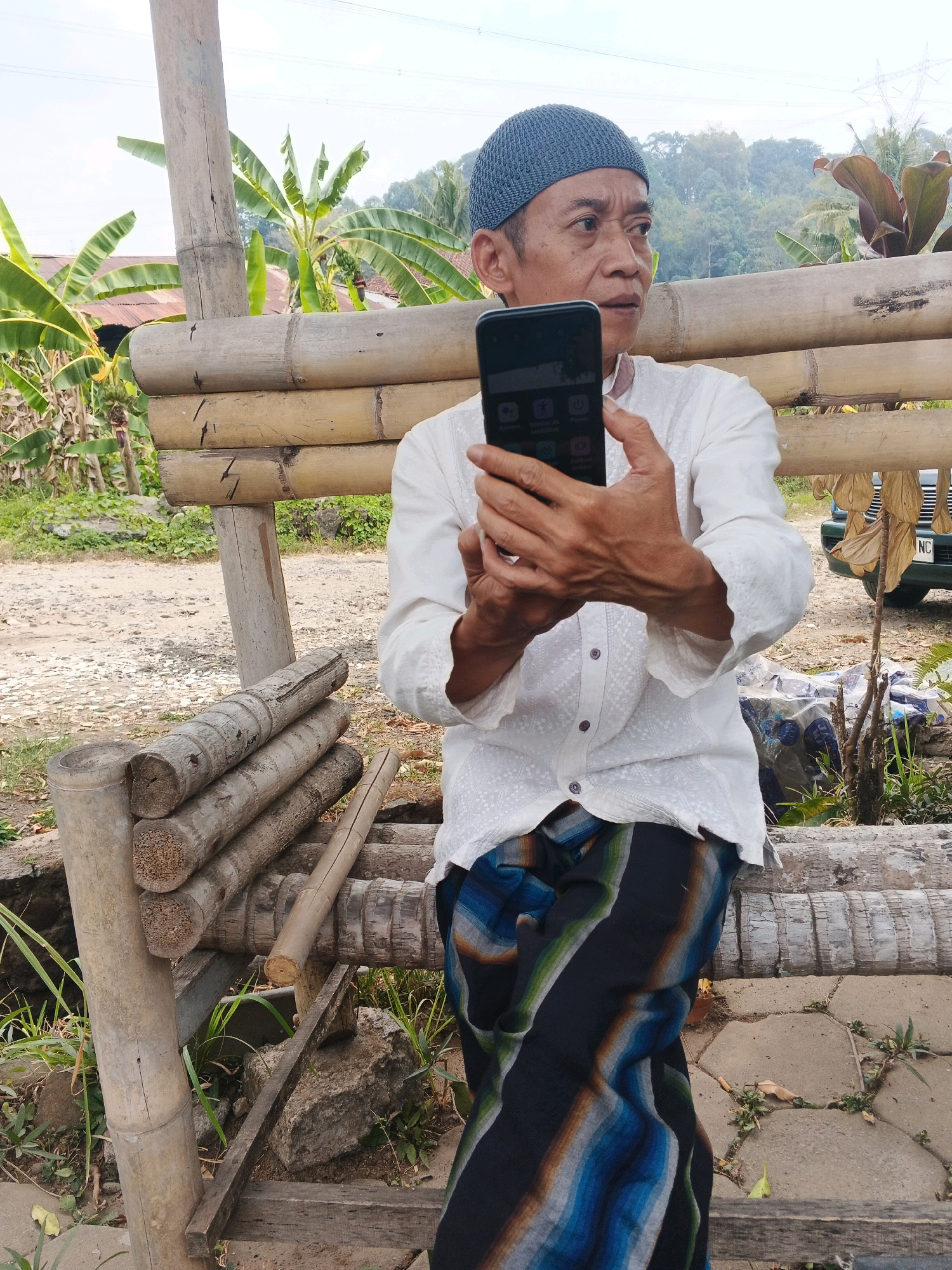 An elderly man wearing a white, button-down shirt with embossed patterns and a blue kufi cap is holding a smartphone in his hands, looking to his right. He is seated on a bench made of bamboo logs, which is part of a rustic outdoor structure. He is also wearing a sarong with vertical stripes of blue, green, and orange. The background shows lush greenery, including banana trees and other tropical foliage, with a slight incline of a hill in the distance. A power line pole and wires are visible in the sky. A green vehicle with a partial license plate reading "NC" is parked to the right of the scene, behind the bamboo structure. The ground is a mix of dirt, gravel, and some paved stones. The scene appears to be in a rural or semi-rural setting, possibly in Ungaran, Indonesia, given the tropical vegetation and architecture.