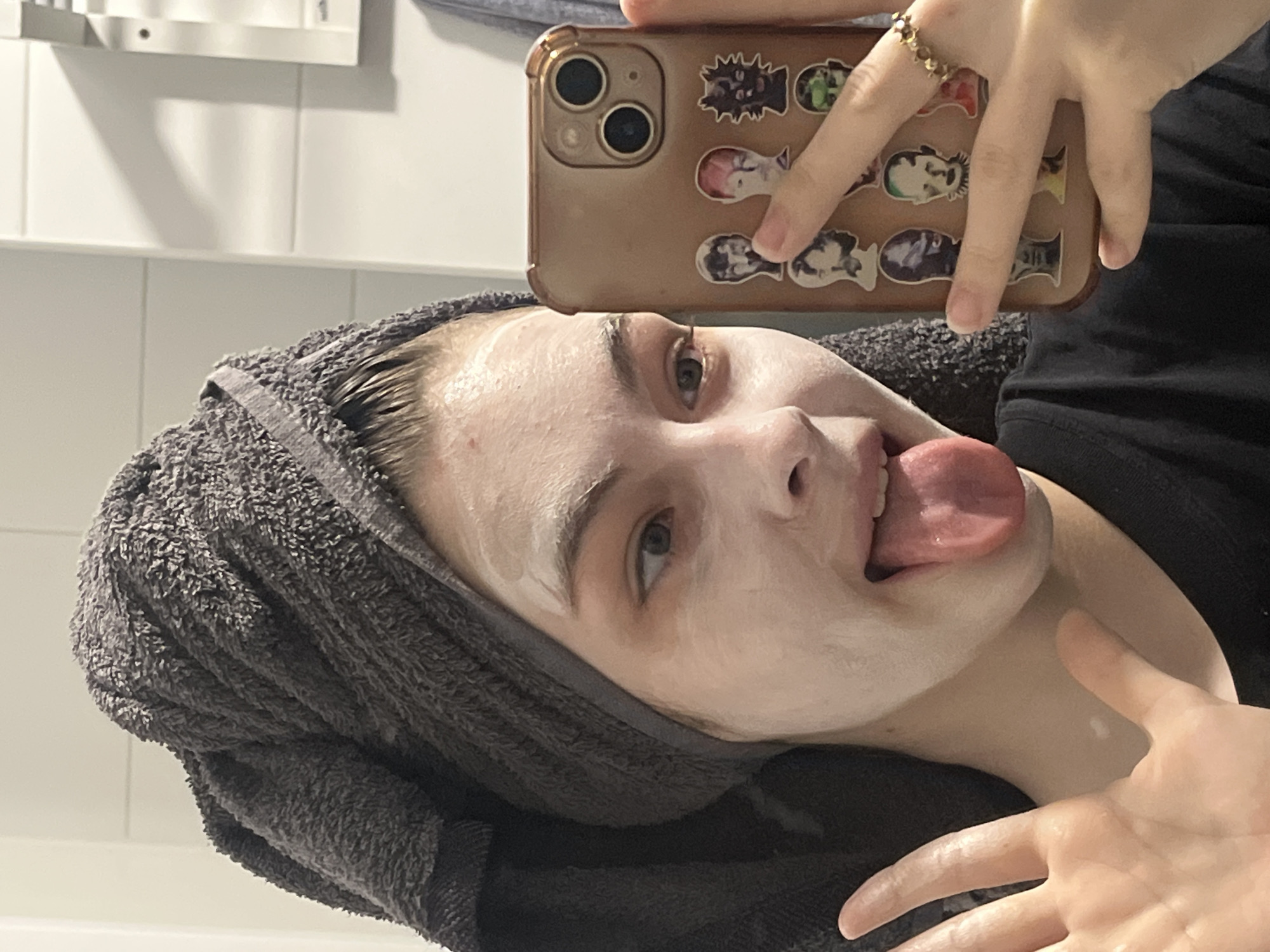 A woman is taking a selfie in what appears to be a bathroom. She has a white face mask on and a gray towel wrapped around her head. Her tongue is sticking out. She is holding a smartphone with a tan case that has stickers on it. She is wearing a gold ring on her finger. The background shows white tiles. The location is not discernible as Saint Petersburg, Russia.