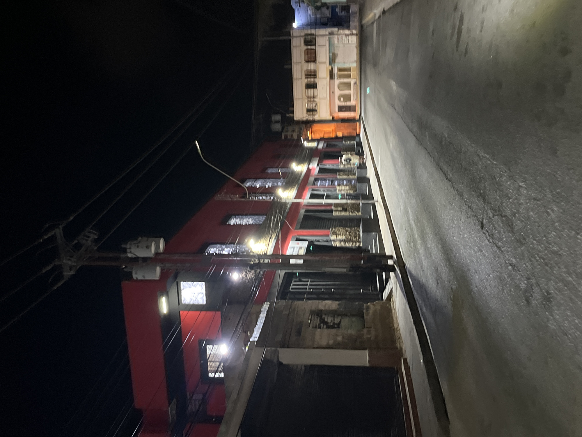 The image depicts a street scene at night in Juan Griego, Venezuela. The street is narrow and appears to be paved with asphalt. The right side of the image shows the road surface with some light reflecting off of it. The left side of the image contains power lines and a red building of several stories. The building appears to have windows with lights illuminated within, and some of the windows are covered with bars. Further up the street, another building is visible. The sky is dark, and the scene appears to be illuminated by streetlights and the lights from the buildings.