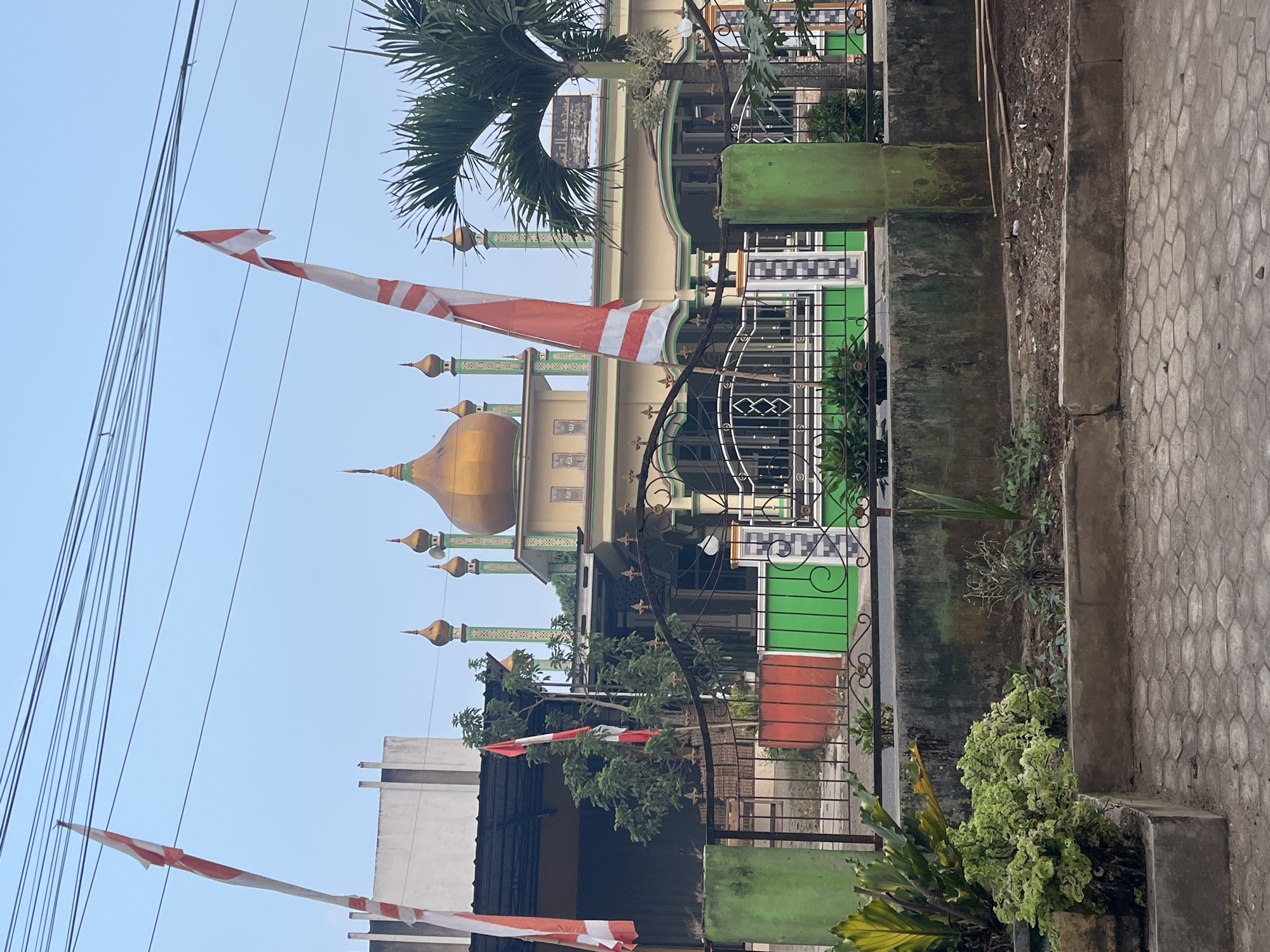 The image depicts a street view in Kemuning, Indonesia, with a prominent mosque as the central subject. The mosque features a golden dome and several green minarets. Two Indonesian flags, red and white, are visible, one flying from a pole on the left and another on a pole near the mosque's entrance. The mosque is enclosed by a black metal fence with green and red decorative elements. Palm trees and other greenery are present, adding to the tropical setting. In the foreground, a paved sidewalk and a low concrete wall with plants are visible. The sky is clear and blue, suggesting daytime. The overall scene is peaceful and captures a moment of local life in Kemuning.