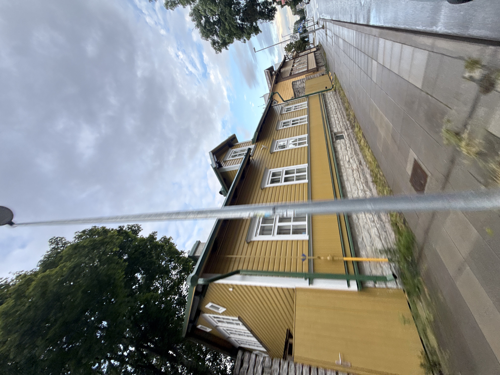 The image captures a view of a yellow wooden house with a green roof and white-trimmed windows, situated on a paved sidewalk in Tallinn, Estonia. A tall, silver street lamp is prominently positioned in the foreground, slightly obscuring the view of the house. The house is constructed with horizontally oriented wooden siding and a stone foundation. Adjacent to the house, a narrow street is visible with a speed limit sign of 30 km/h, indicating a residential or commercial area. The sky is overcast with clouds, and trees line the background. The wet pavement suggests recent rainfall.