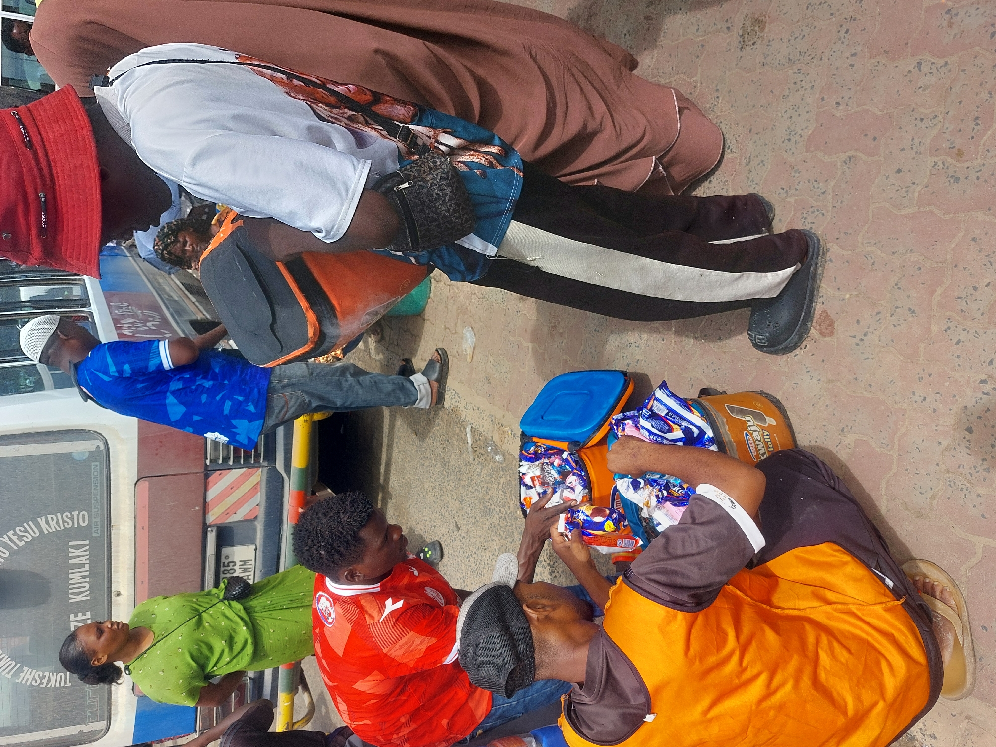 This medium-angle, high-perspective shot depicts an outdoor gathering in Dar es-Salaam, Tanzania, under bright daylight. Several individuals are present, standing and seated on a paved surface.
In the foreground, two men are seated on the ground. One man wears a red t-shirt, likely a football jersey, and blue jeans. Beside him, another man, dressed in an orange and brown t-shirt with dark pants and sandals, is reaching into a blue cooler bag. The bag is filled with various packaged snacks, including items like "Chupa Chups," "Milky," and "SWEET JOY Jelly," suggesting distribution or consumption of treats.
Behind the seated men, closer to a parked white and brown bus, a woman in a green dress and a man in a blue shirt with a white skullcap (kufi) are standing. The bus features visible text on its window: "NDIO YESU KRISTO" (Yes, Jesus Christ) and "TUKESHE TUMLAKI" (Let's wake up to meet Him), with "AIR SUSPENSION" also visible. The bus license plate reads "385 AWM".
To the left, a person wearing a red bucket hat, a white t-shirt, and black pants with a white stripe stands, carrying a small patterned shoulder bag. Another person in a long brown garment is partially visible behind them. The ground is paved with light-colored interlocking concrete blocks. The scene appears to be a casual public space, possibly a bus stop or market area.