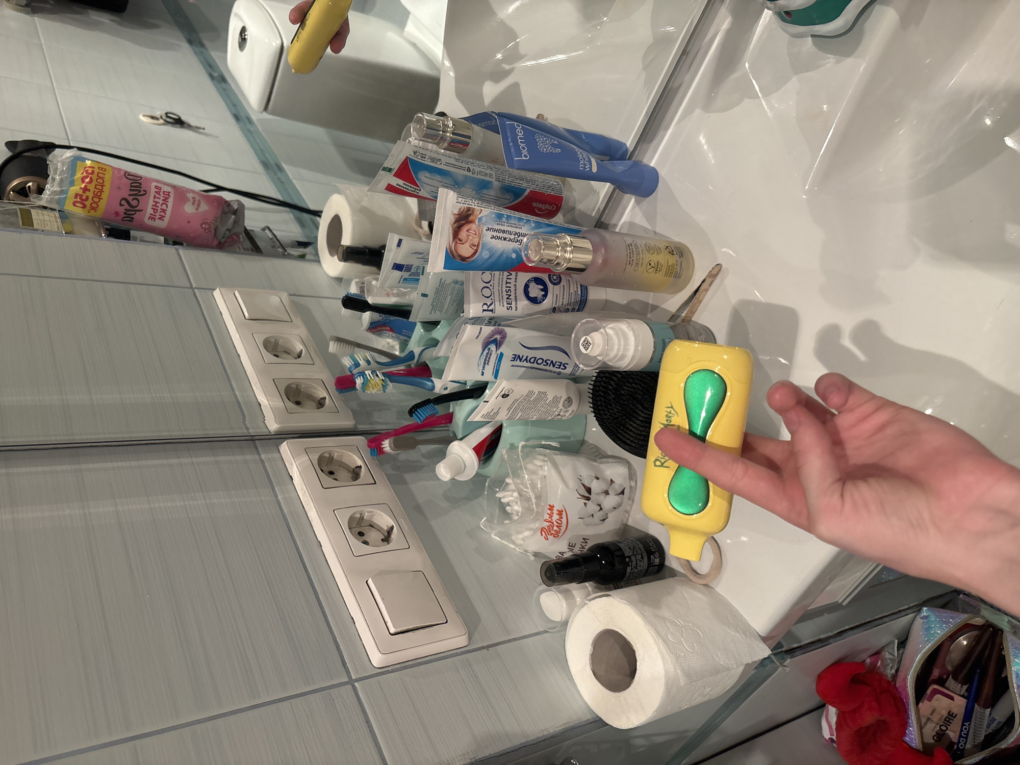 The image displays a bathroom sink area with various toiletries and personal care items. On the sink counter, there is a roll of toilet paper, a black spray bottle, a bag of cotton swabs, and several tubes of toothpaste and other cosmetic products. A hand is visible in the foreground, holding a yellow and green item. A mirror reflects the scene, showing more toiletries and a toilet in the background. The wall tiles are light blue. The specific location in Sinyavino, Russia cannot be determined from the image alone.