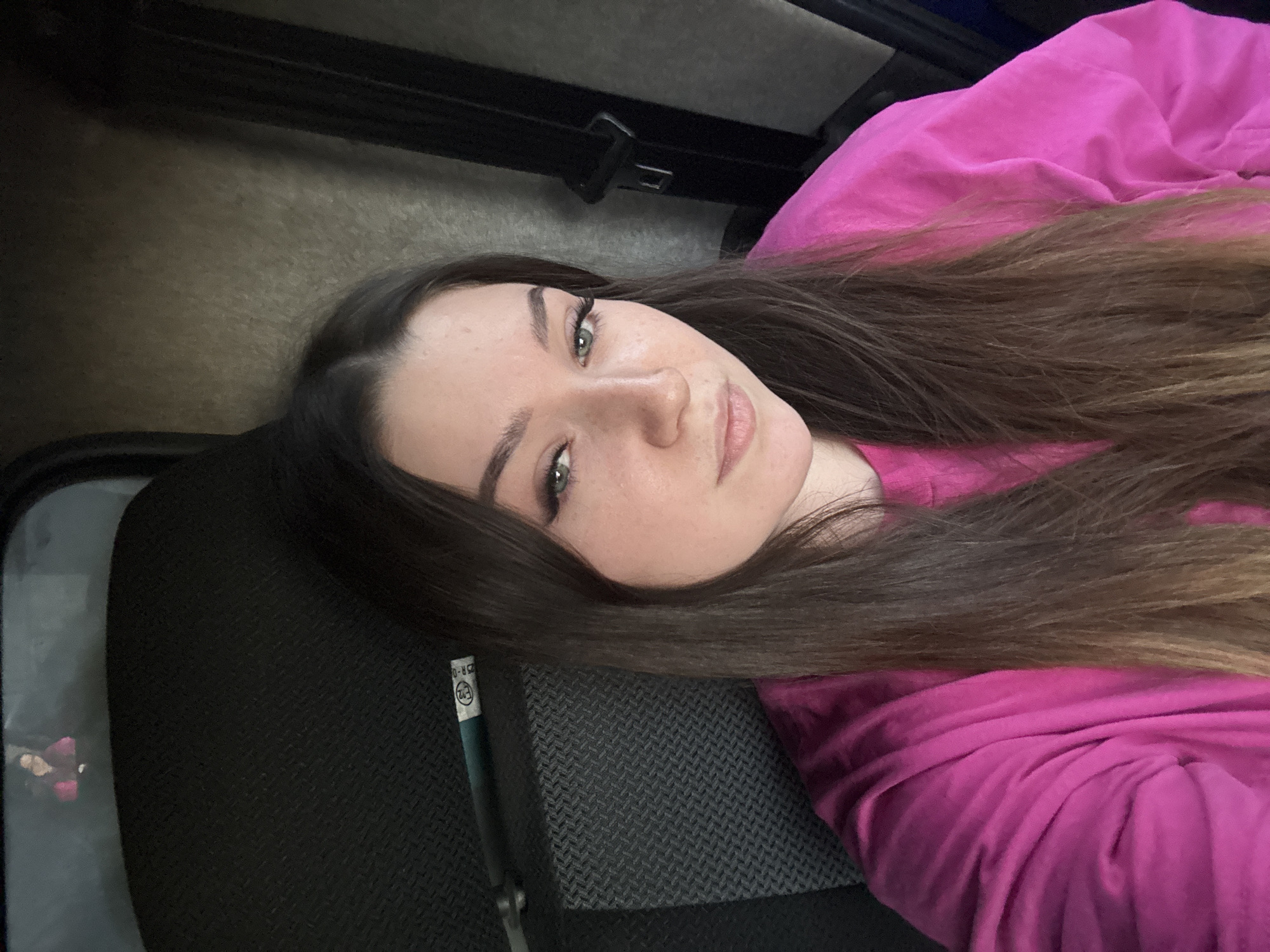 The image is a selfie of a young woman with long, dark brown hair, wearing a bright pink sweatshirt. She is sitting in what appears to be a vehicle, possibly a bus or truck, as indicated by the seat texture and the seatbelt visible on the right side of the frame. The woman is looking directly at the camera with a neutral expression. In the reflection of the upper window, a smaller image of the woman wearing a pink top can be seen. The environment is dimly lit. The location is not specified to be Krasnokamsk, Russia.