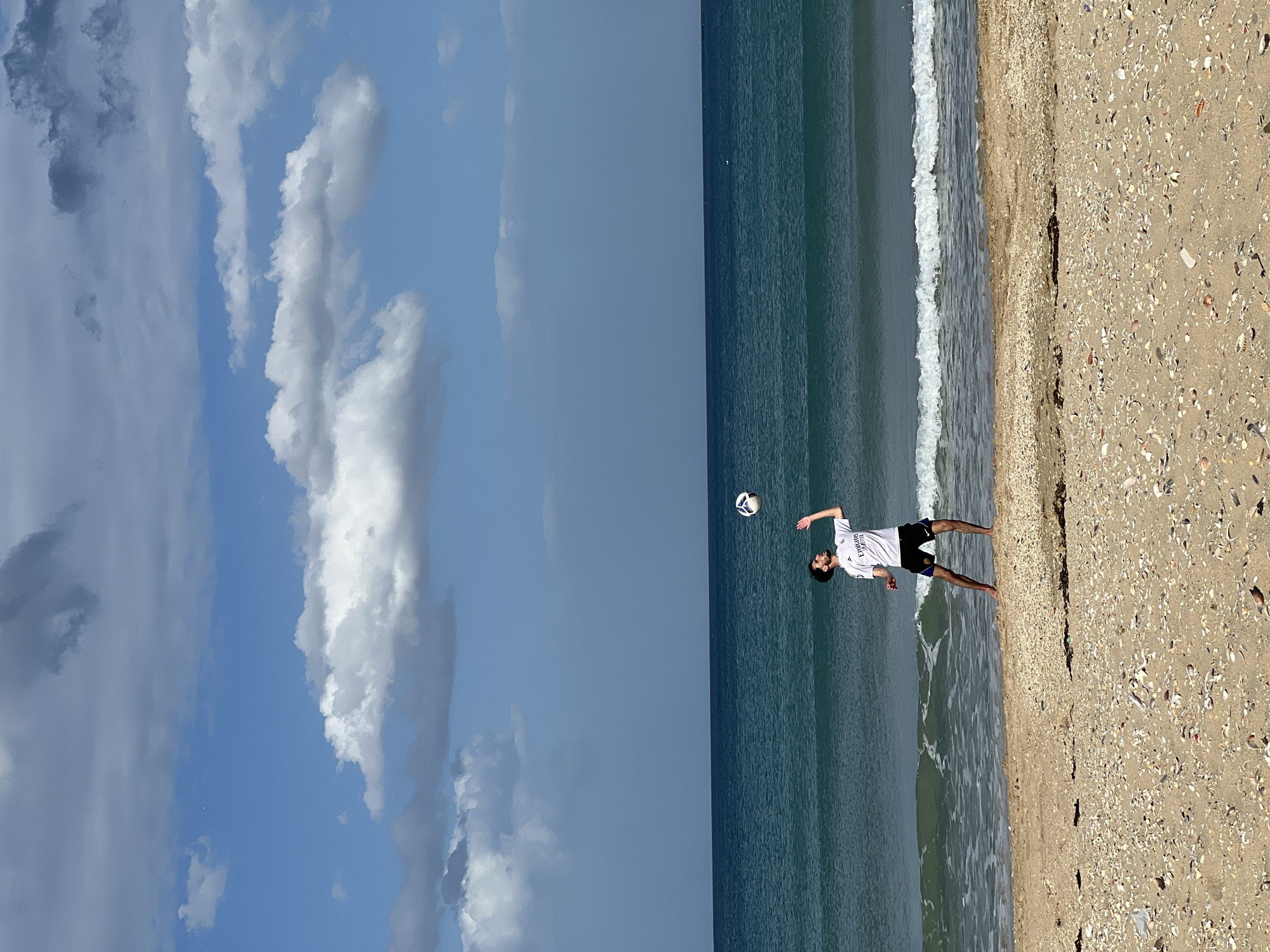 A man in a white t-shirt and black shorts stands on a beach in M'Diq, Morocco, facing the sea. He is throwing a soccer ball. The beach is sandy with shells. The sea is calm with small waves. The sky is partly cloudy.