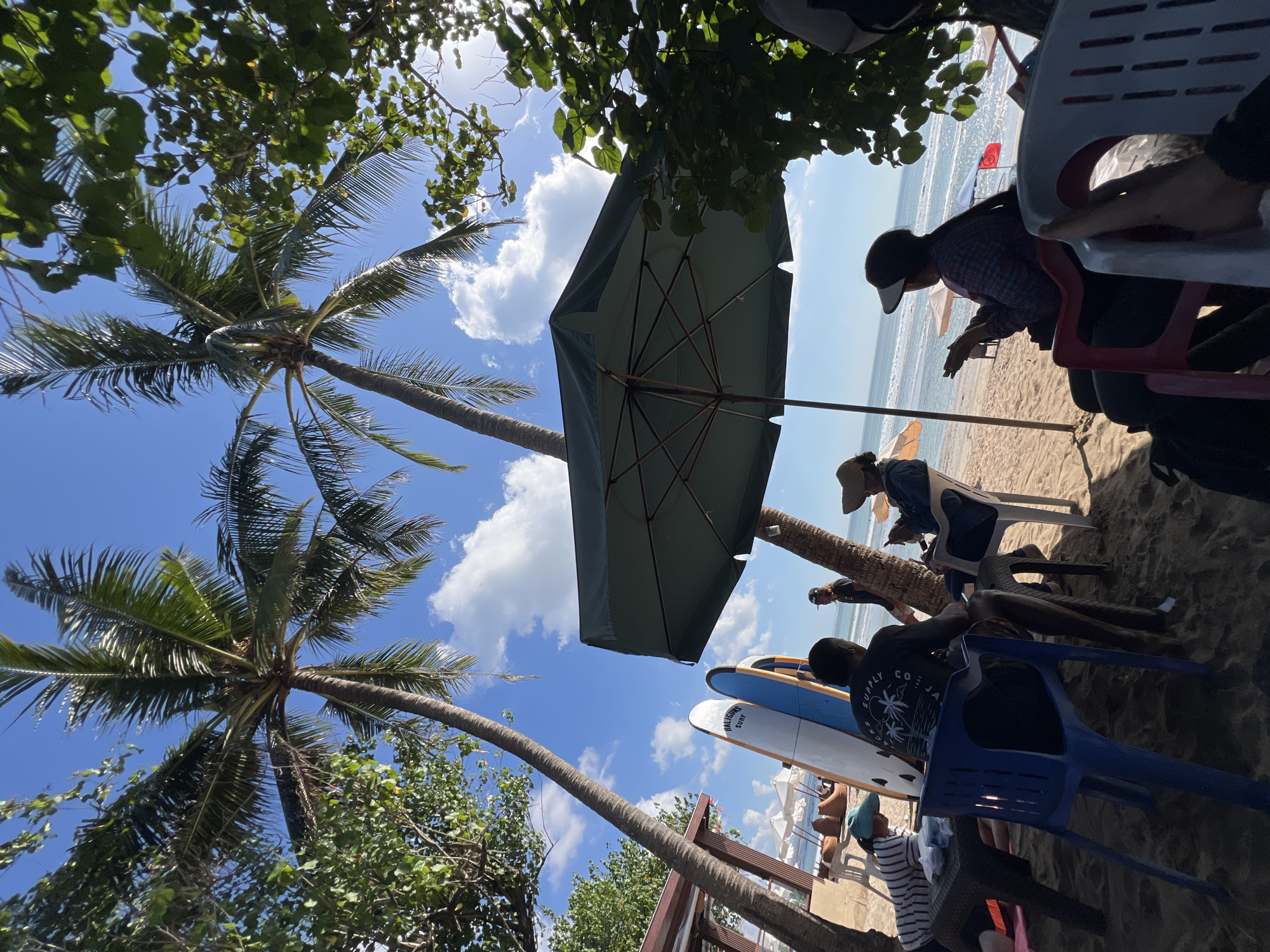 In Kuta, Indonesia, a beach scene unfolds under a bright, partly cloudy sky. Two palm trees with lush fronds cast shadows on the sandy shore where several people are gathered. In the foreground, an olive green beach umbrella provides shade.
To the left, three surfboards stand upright against a wooden structure, one white and two blue, suggesting a surf spot. A table with a dark brown top is positioned next to them, with a person in a striped shirt and a teal hat sitting on a blue chair. Another person, wearing a black t-shirt with a palm tree graphic, is seated on a blue plastic chair facing away from the camera, with their legs visible. A third individual, wearing a denim jacket and a hat, sits on a white plastic chair to the right, looking at their phone.
In the background, the ocean stretches out, with gentle waves and a few scattered beach umbrellas on the sand. The overall atmosphere appears relaxed, with people enjoying a day at the beach.