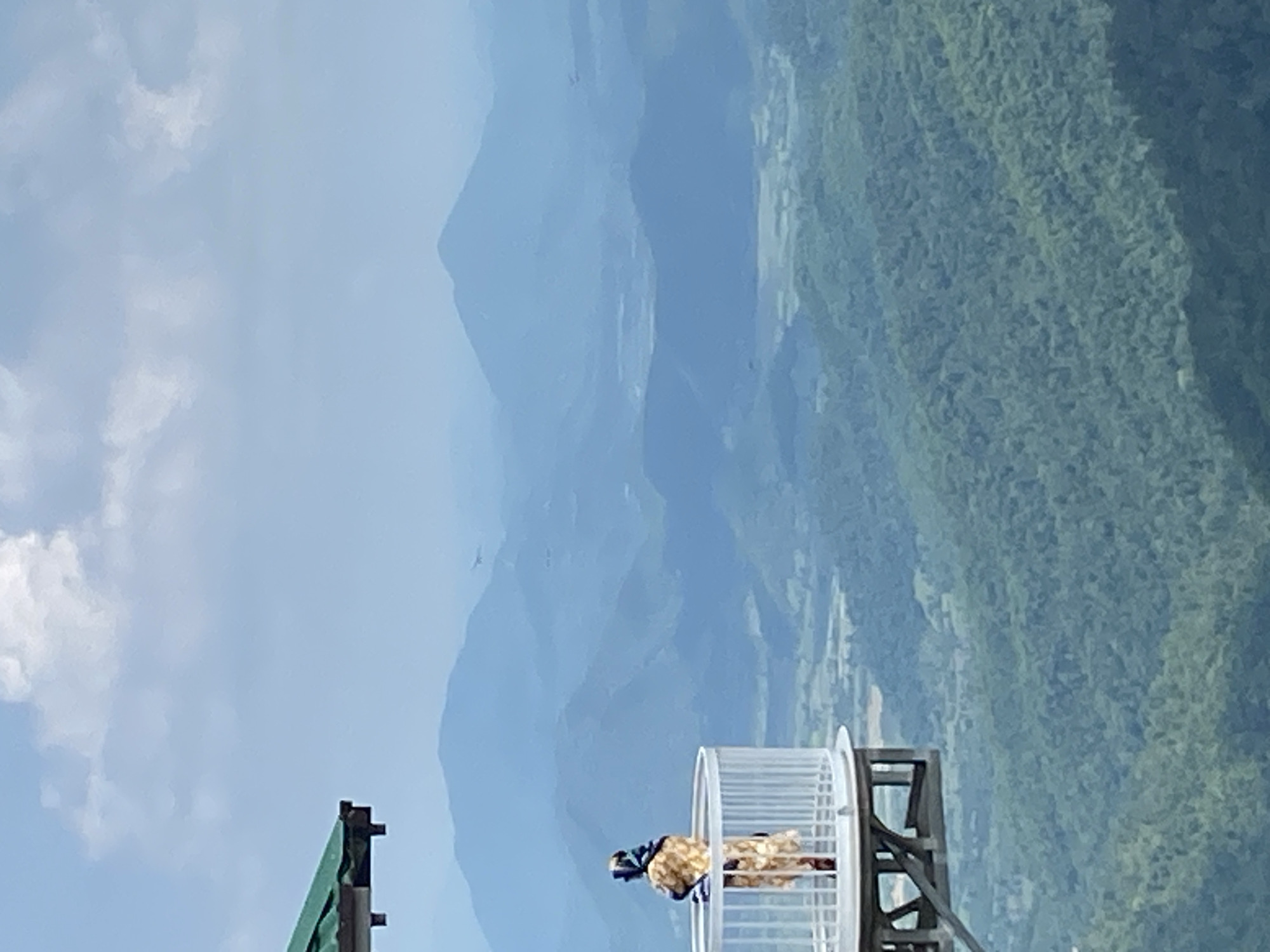 A person stands on a white, circular observation deck, looking out at a vast expanse of mountains. The person is wearing a light-colored dress with a patterned design and a headscarf. The observation deck is supported by a wooden structure and has a railing. A portion of a green-roofed structure is visible in the upper left corner. The background is a series of layered, hazy blue mountains, with the closest ones covered in dense green forest. The sky is a pale blue with scattered white clouds. The scene appears to be at a high elevation, offering a panoramic view of the natural landscape. The location is identified as the observatory in Nam Hải, Vietnam.