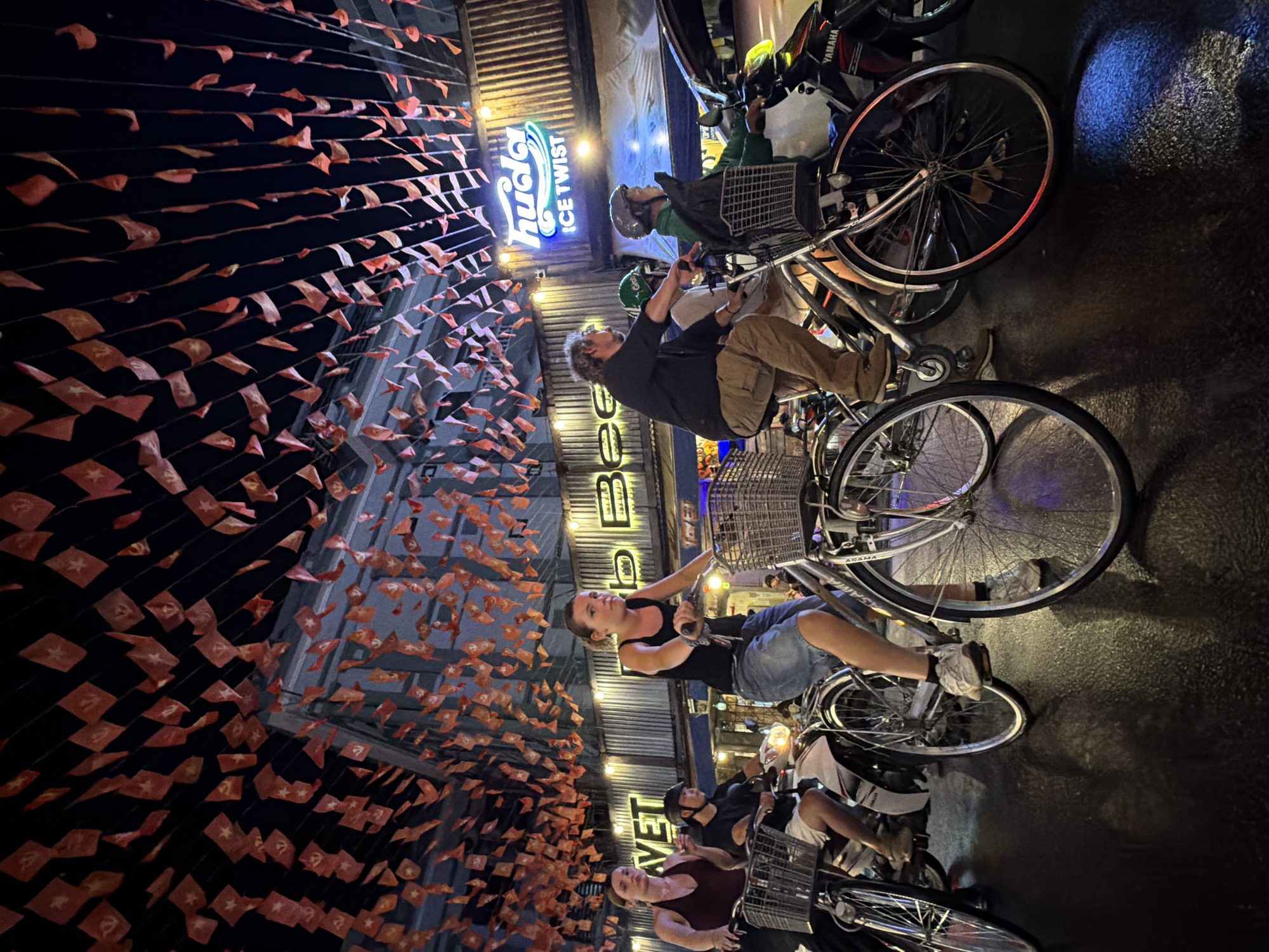 The image captures a street scene at night in Thuận Hóa Ward, Vietnam. The foreground is dominated by the wheels and lower bodies of several bicycles and a scooter, suggesting movement or a pause in transit. Several individuals are visible, some riding bicycles and others on a scooter. The people are dressed casually in tops and shorts or skirts. They appear to be navigating the street, with some looking forward and others towards the camera.
The background features a building with a corrugated metal facade, illuminated by neon signs. One sign displays the word "huda" in a stylized font above "ICE TWIST," indicating a beverage brand. Another sign reads "BEER." Above the street, a dense canopy of small, red, triangular flags is strung across, creating a festive or decorative atmosphere against the dark night sky. The street surface appears wet, reflecting the ambient light and creating glossy patches. The overall scene depicts a bustling urban environment at night, with a blend of pedestrian and vehicular traffic under a canopy of flags.